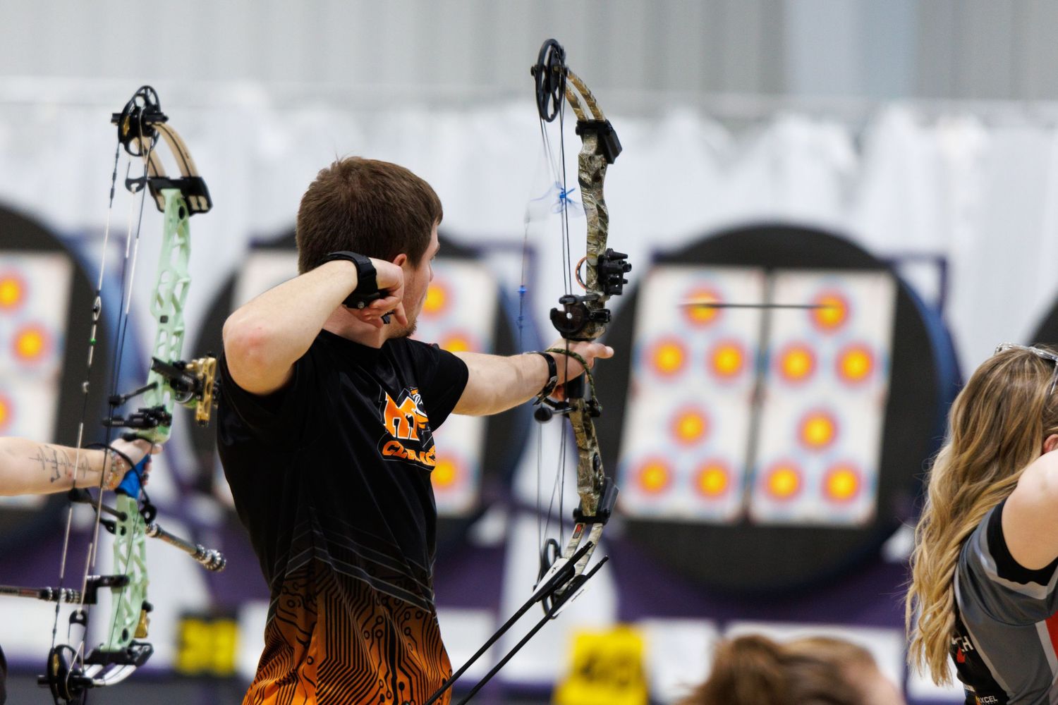 Young male archer drawing a compound bow at an indoor archery competition with colorful targets behind him.