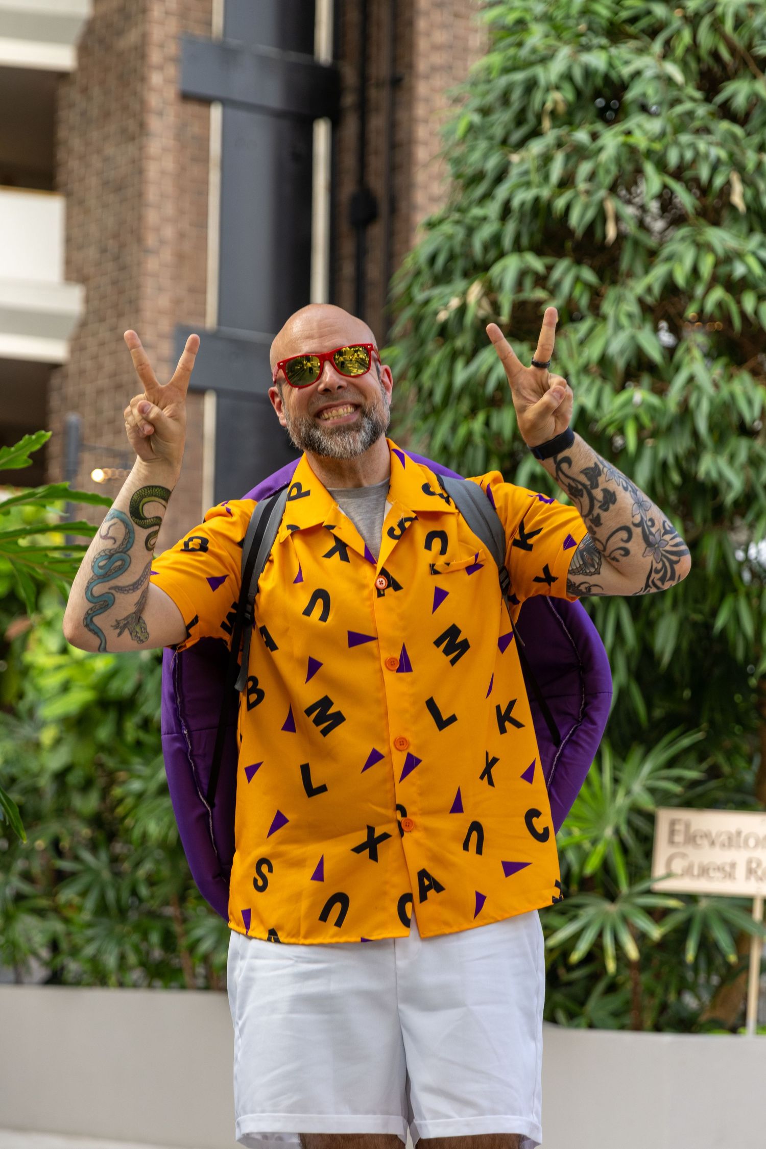 Someone wearing bright yellow patterned shirt and white shorts makes peace signs while standing outside brick building.