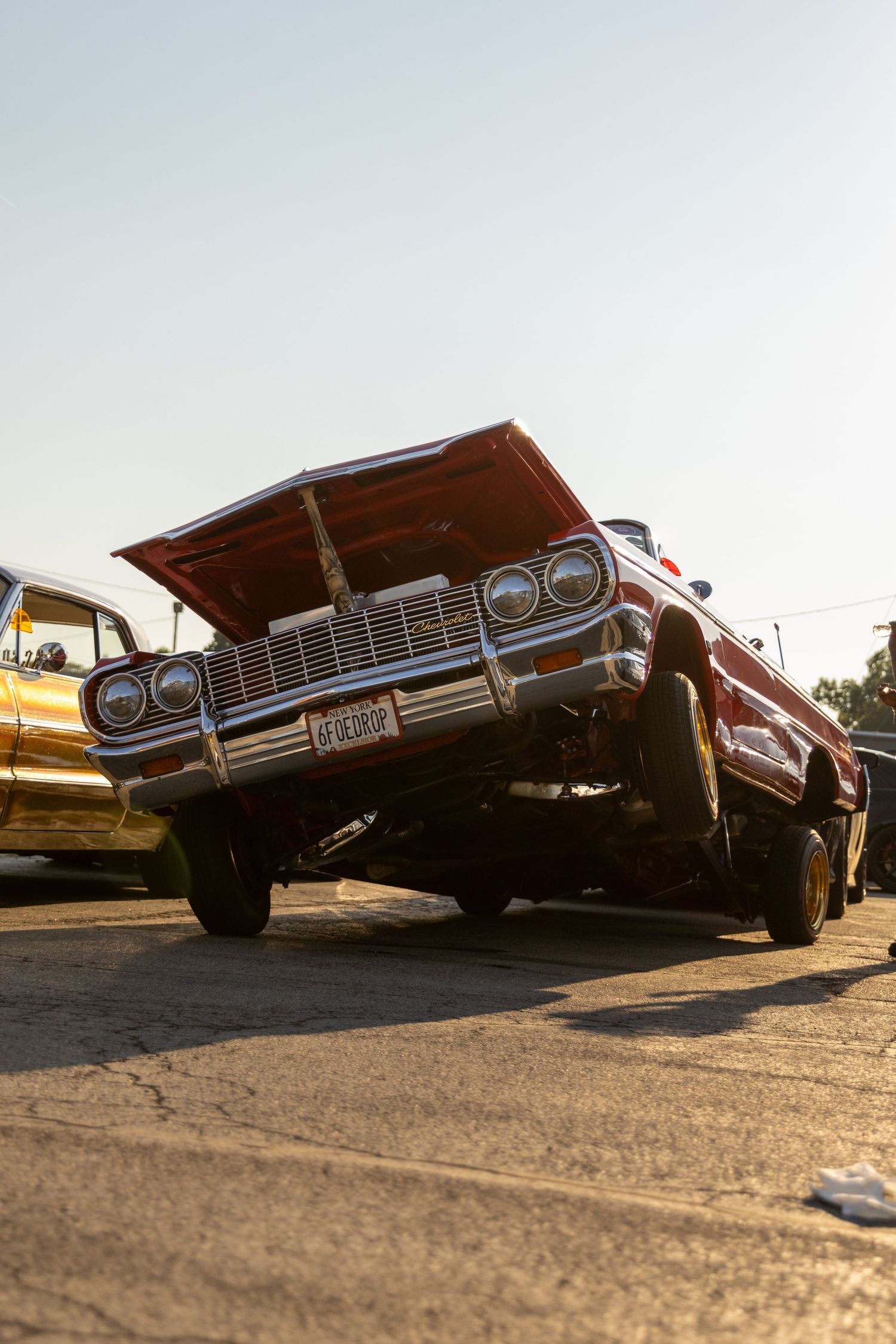 A classic red Chevrolet lowrider car with hydraulics lifted at an angle on the street during golden hour.