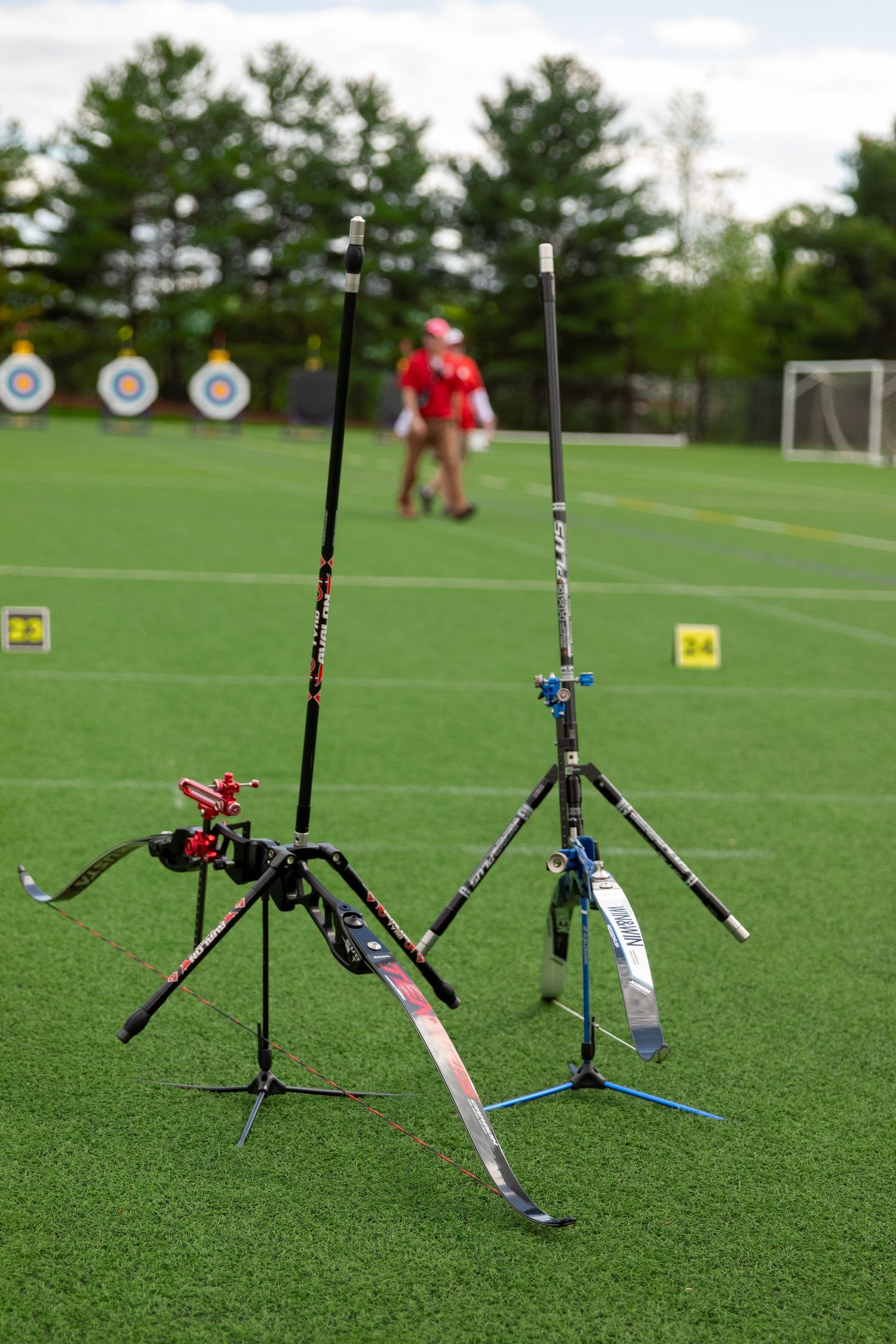 Two archery bow stands set up on green field with target practice equipment in background.