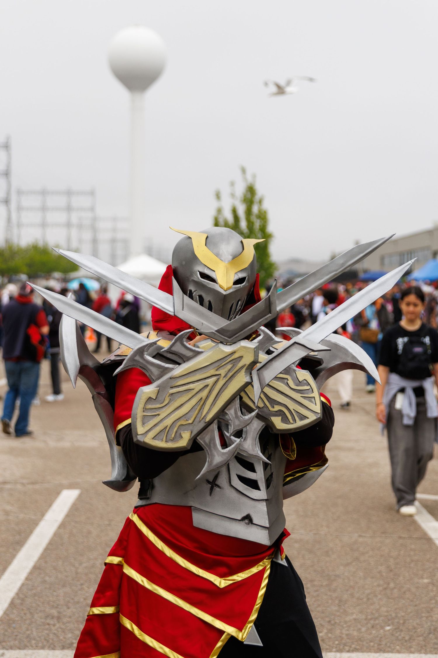 Cosplayer in detailed red and silver armored knight costume crosses dual swords at outdoor comic convention.