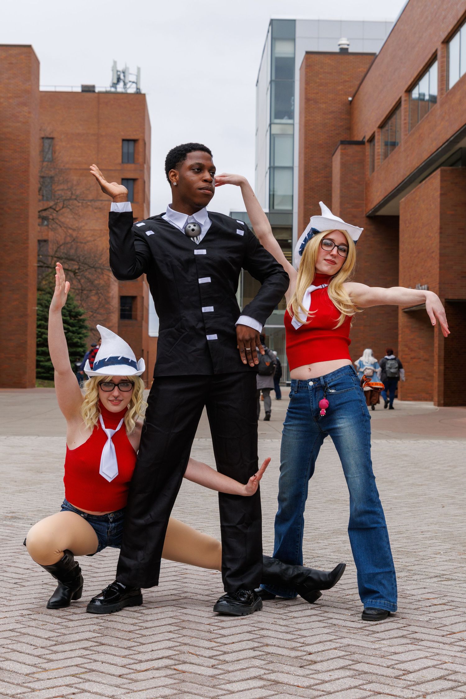 Three cosplayers pose dramatically outdoors near brick buildings, two blonde women in red tops and one tall man in black uniform.