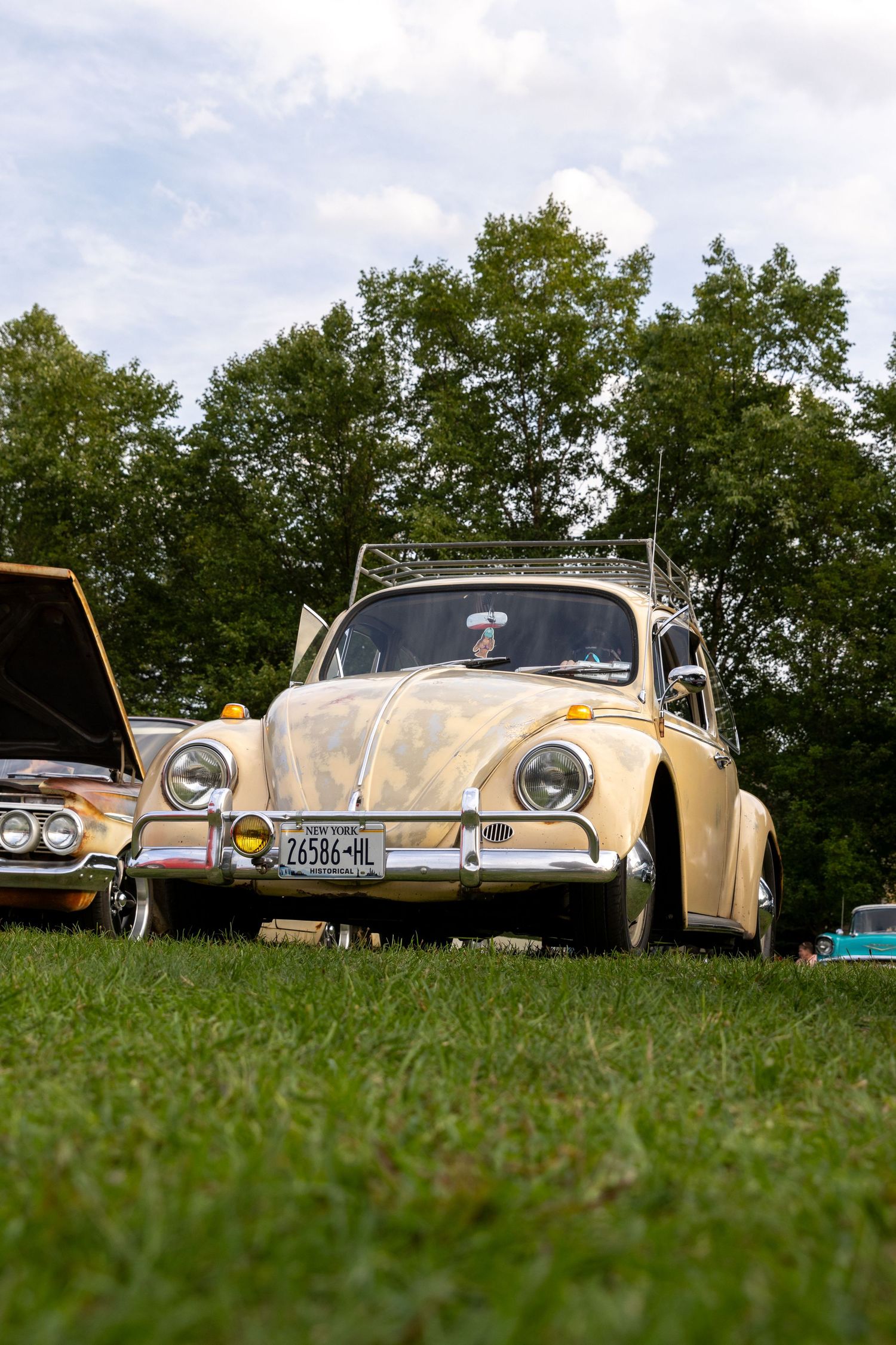 A vintage cream-colored Volkswagen Beetle parked on grass with trees in the background on a cloudy day.