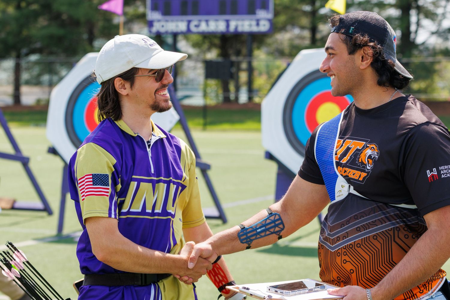 Two archers in competition gear shake hands at an outdoor archery range with colorful target faces in the background.