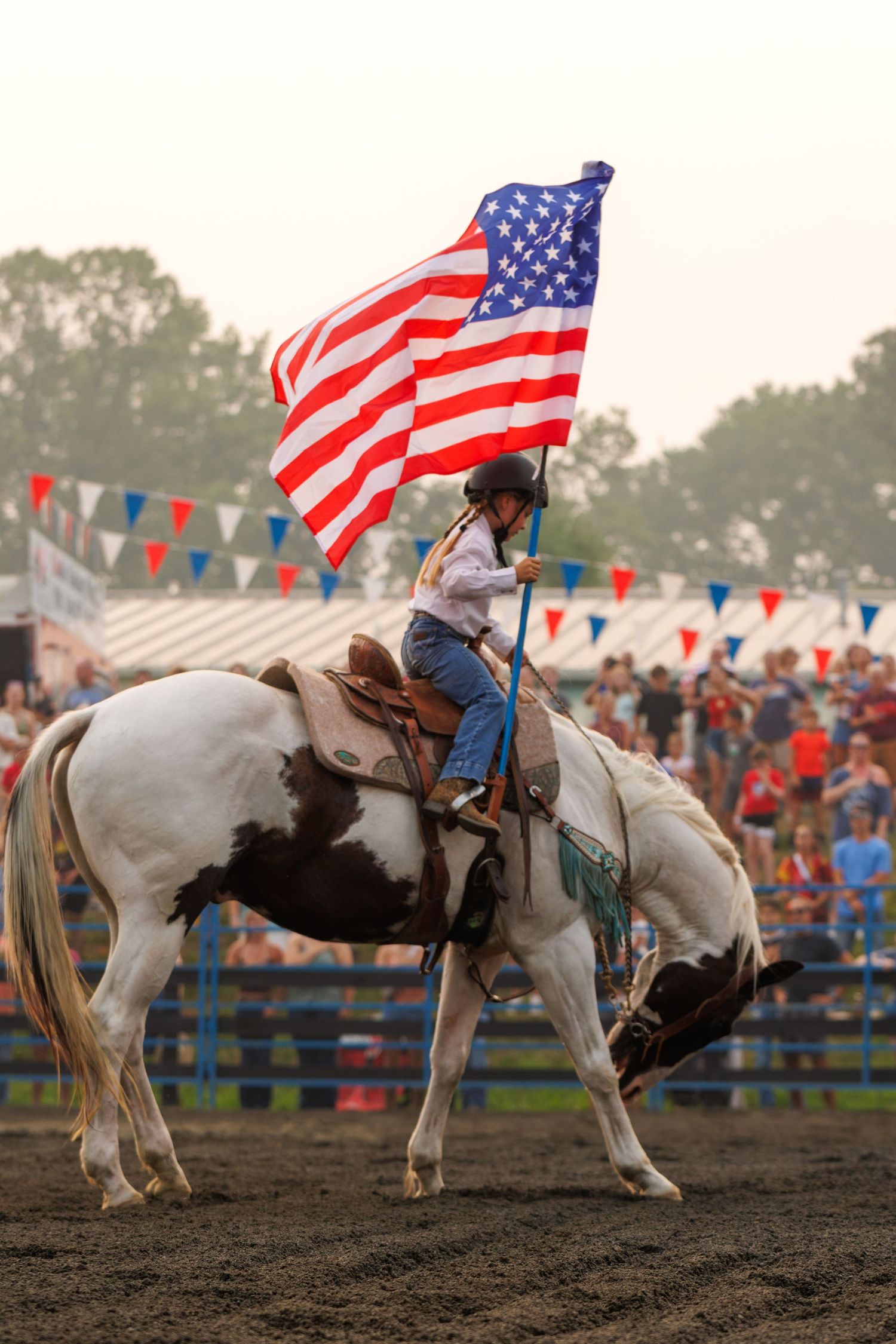 Rider on a paint horse carries a large American flag at a rodeo arena with colorful bunting in the background.