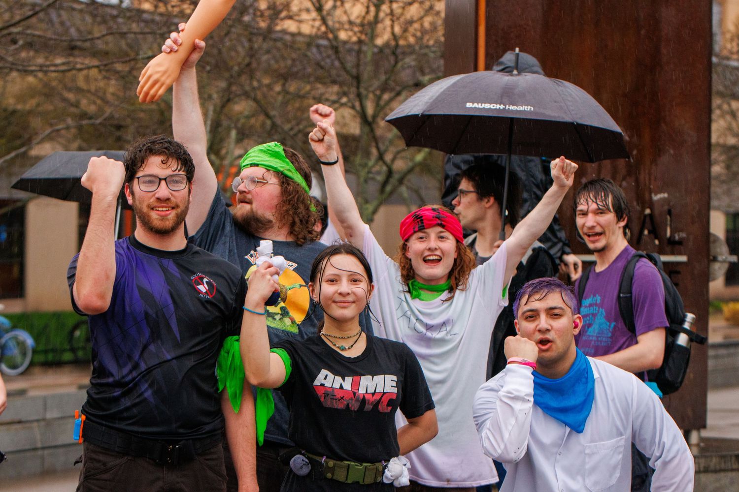 Group of cosplayers posing enthusiastically outdoors in the rain, one holding an umbrella, wearing colorful costumes.