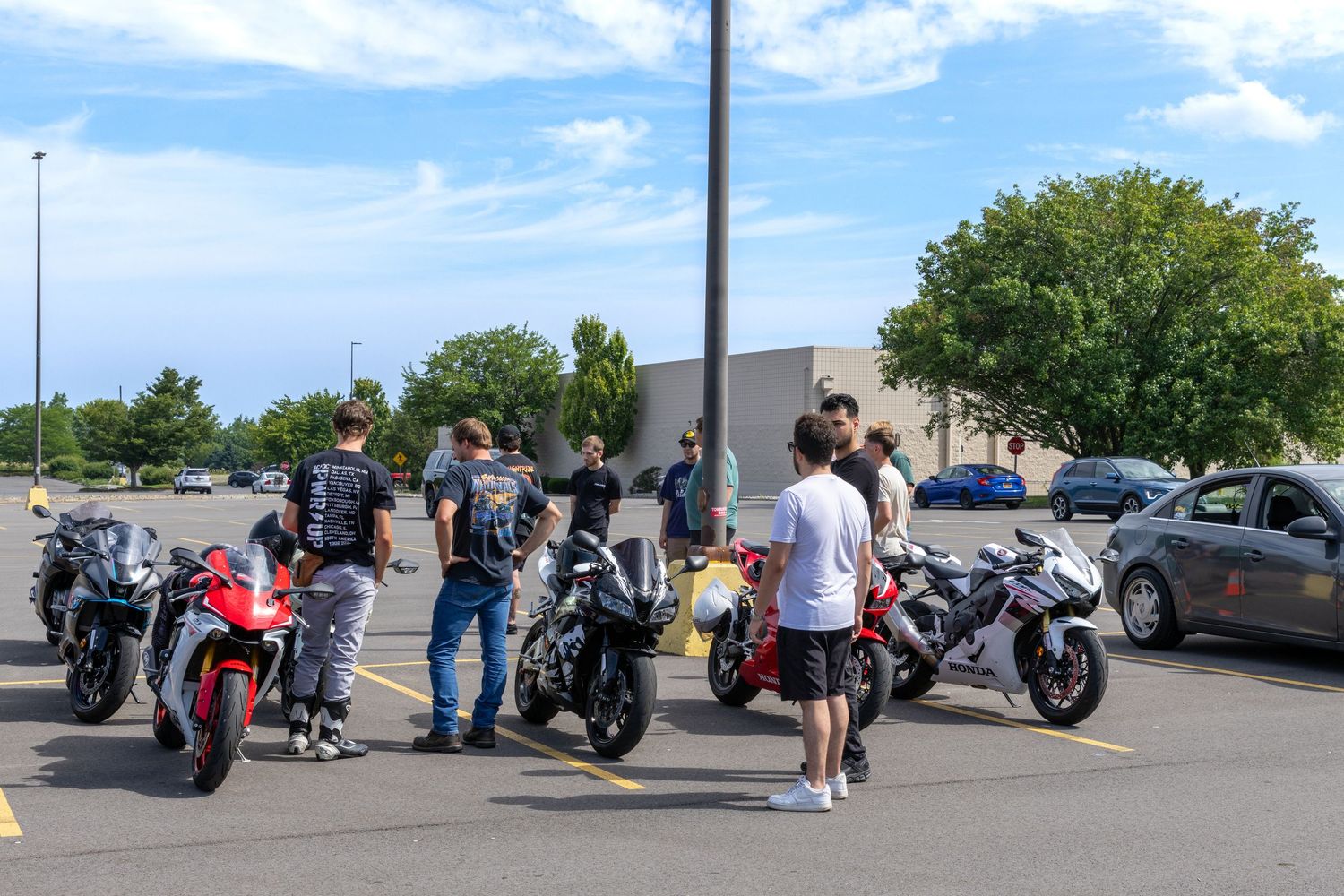 Motorcycles lined up in a parking lot with riders gathering under blue skies on a sunny day.