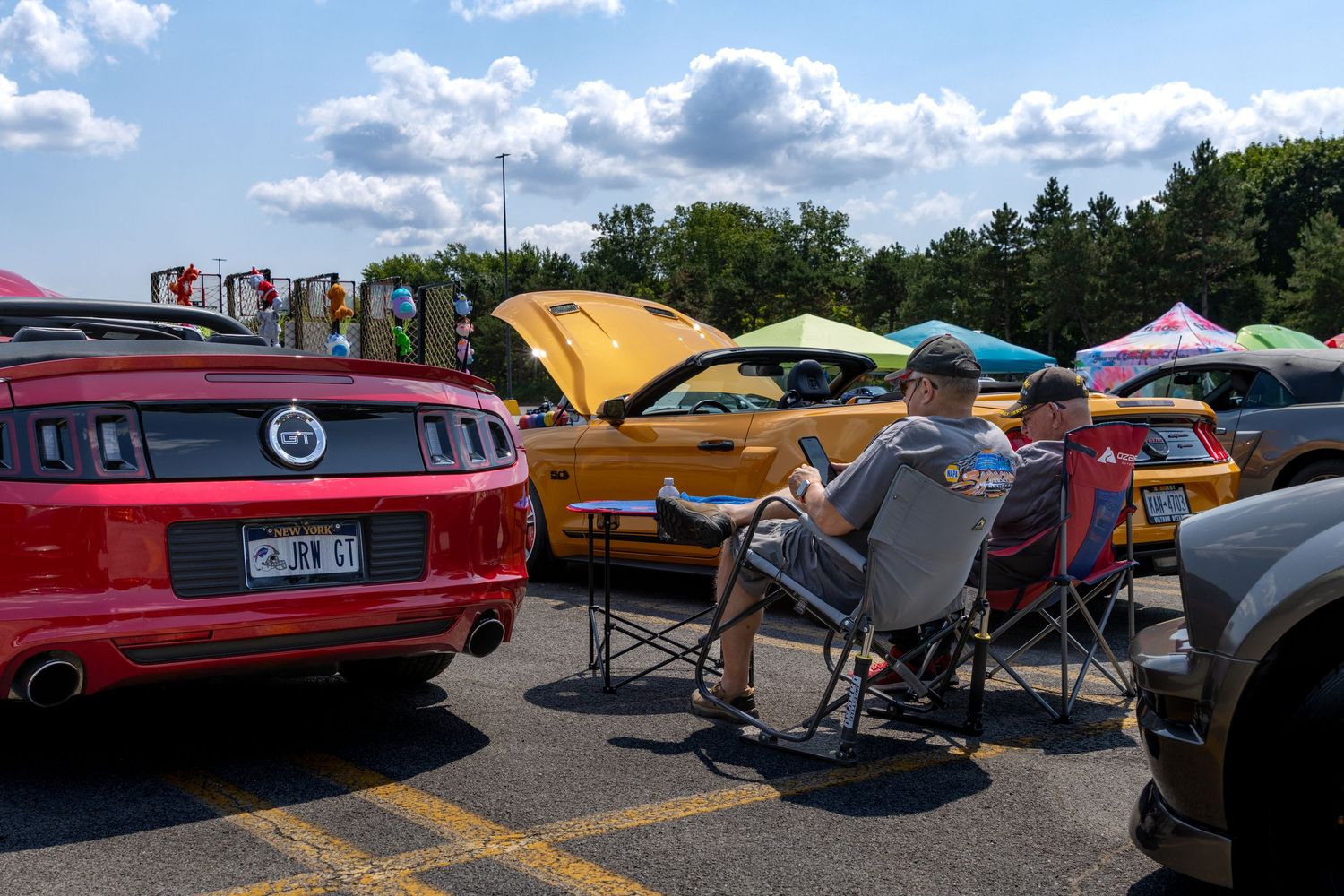 People relax in camp chairs at a car show with Ford Mustangs and other vehicles on display under blue summer skies.