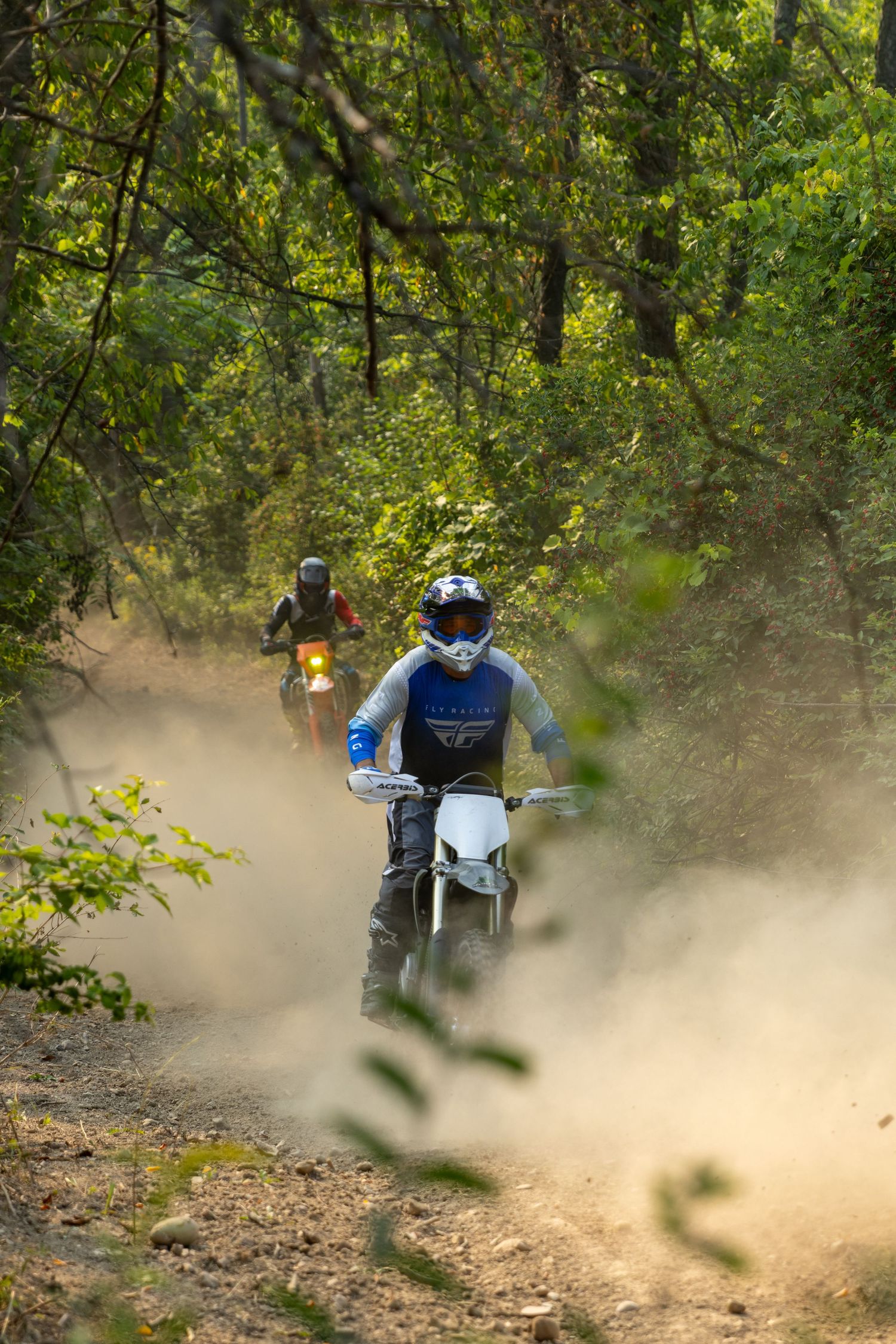 Motorcyclists riding through a wooded trail creating clouds of dust as they navigate the dirt path.