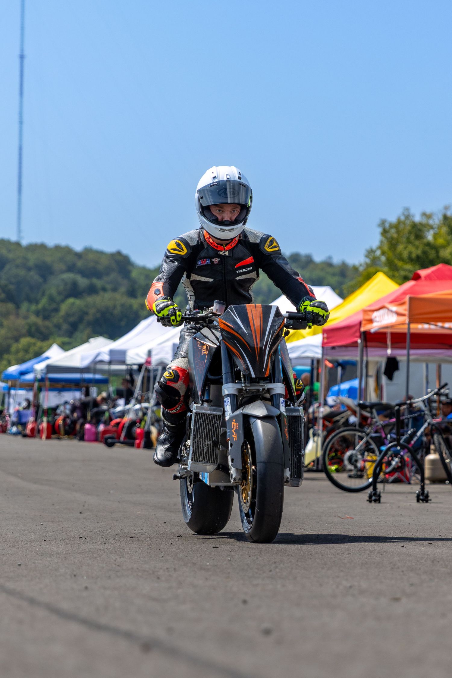 A ground-level shot shows a lineup of custom motorcycles parked at an outdoor event with colorful vendor tents in background.
