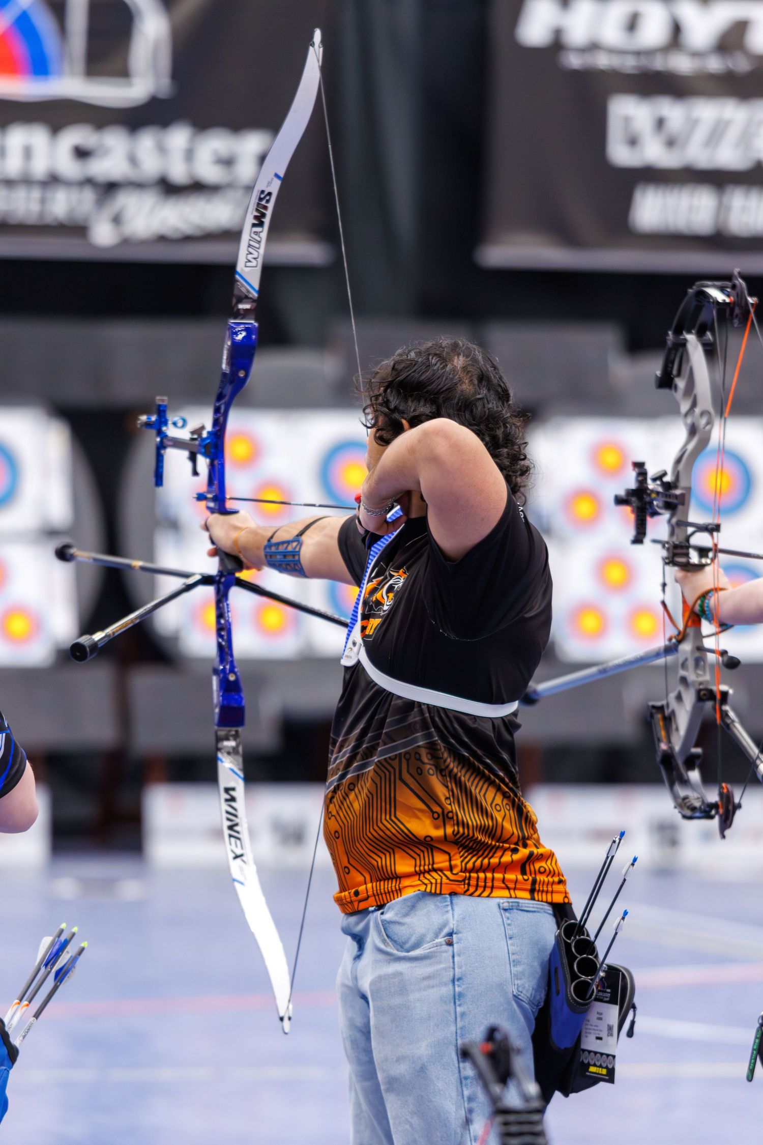 Archer drawing a recurve bow at an indoor archery competition, with targets visible in the background.