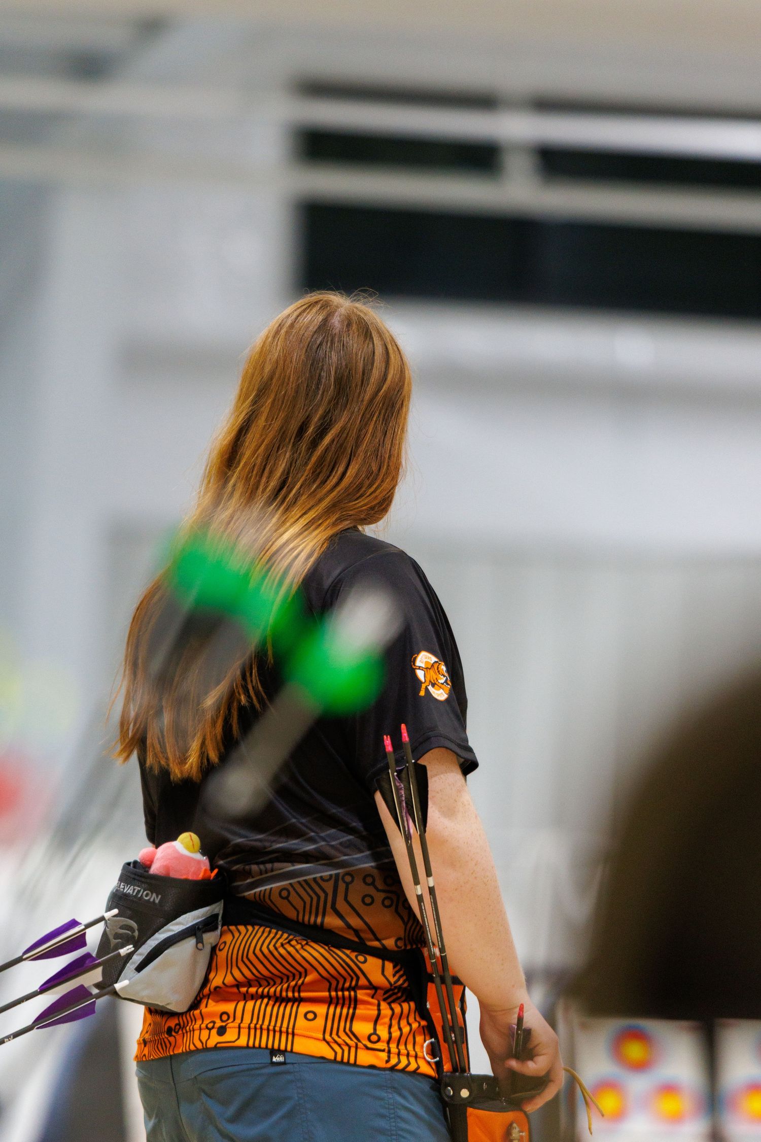 Female archer seen from behind holding a bow with arrows in quiver at an indoor archery competition.