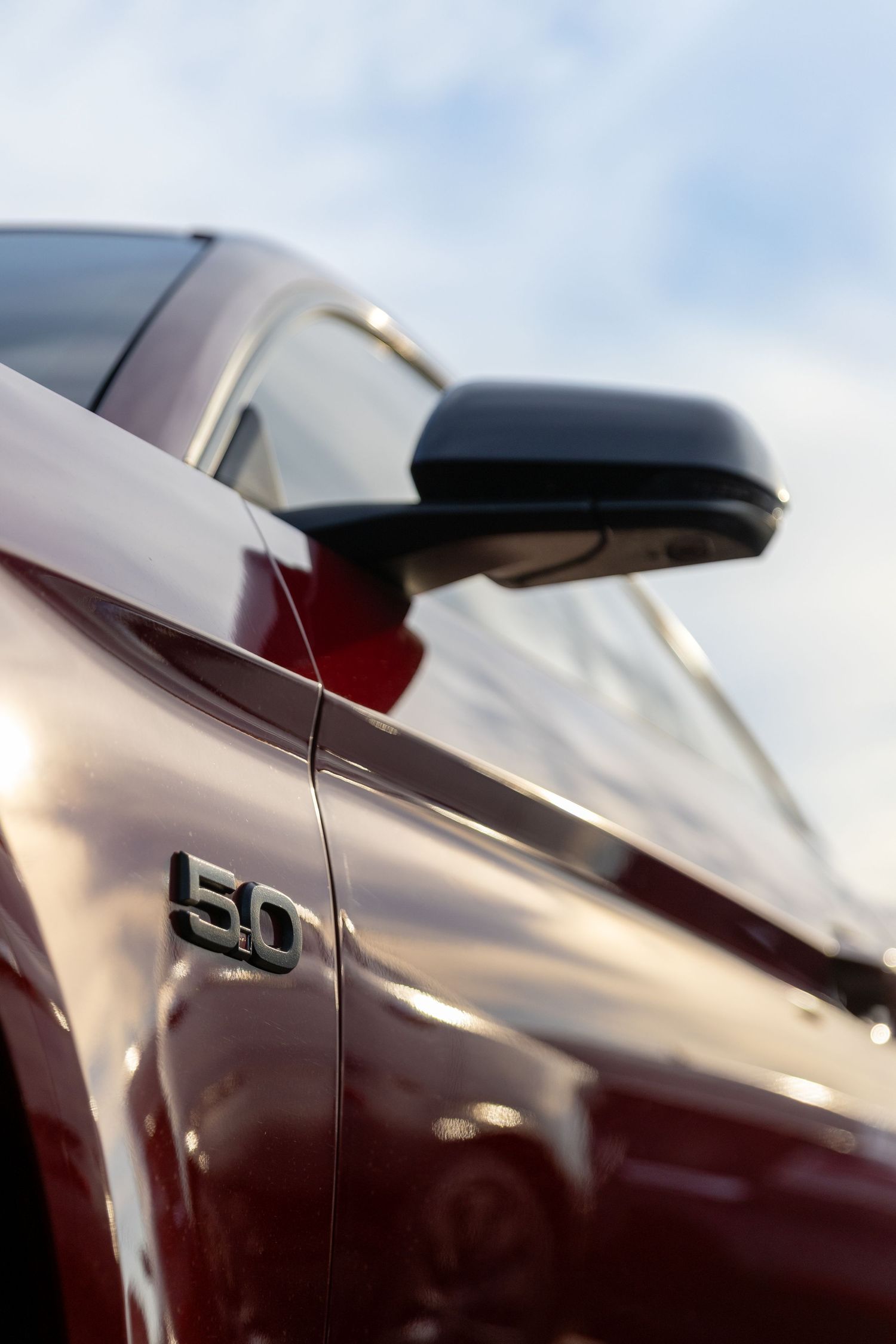 Close-up detail of a maroon car's side mirror and 5.0 badge emblem against a soft blue sky background.