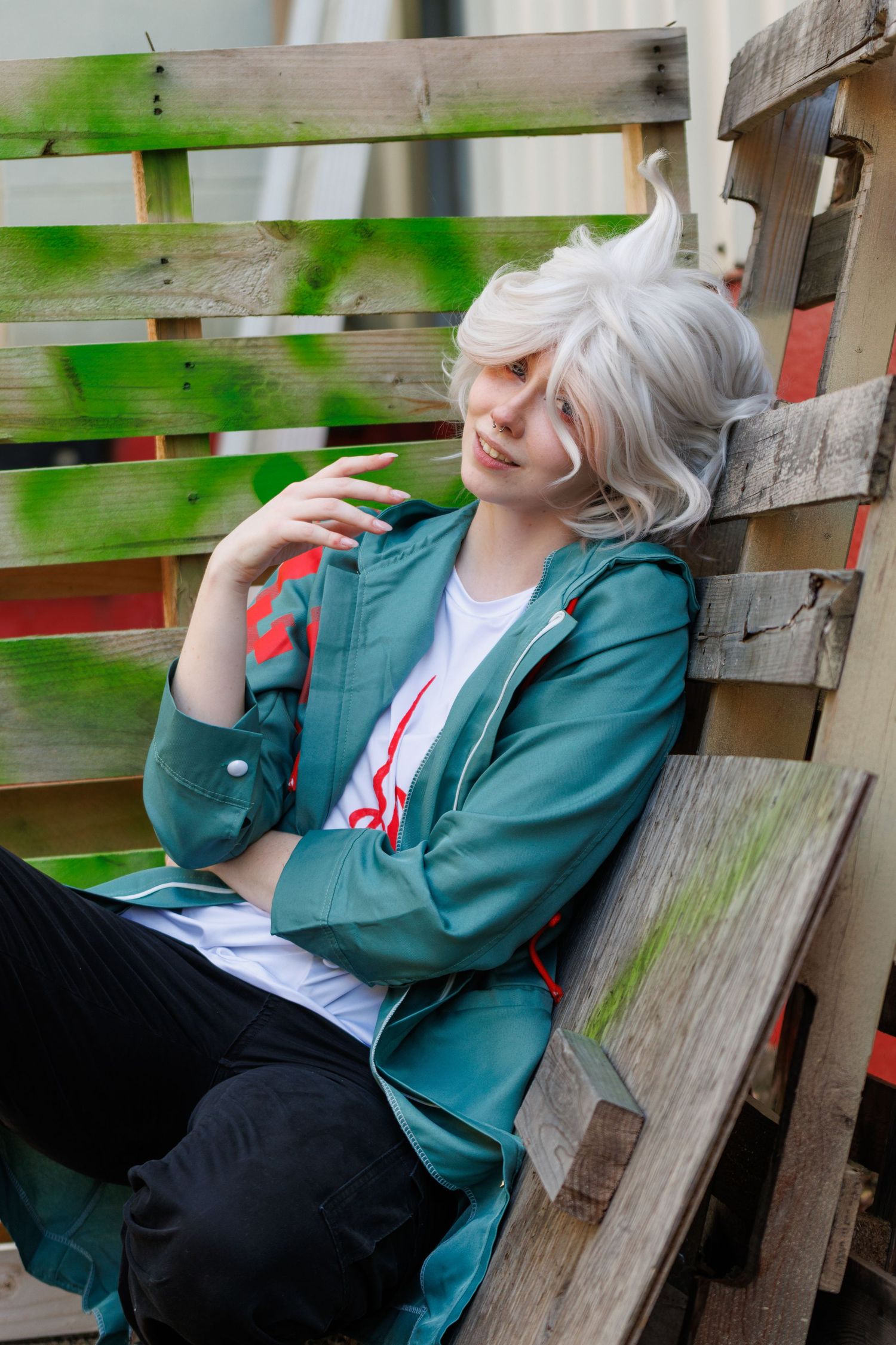 Cosplayer with fluffy white hair in a teal jacket and black pants leans back on a weathered green wooden pallet bench.