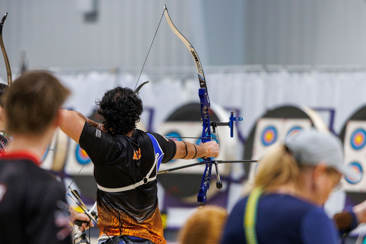 Archer drawing recurve bow at colorful targets in indoor archery tournament.