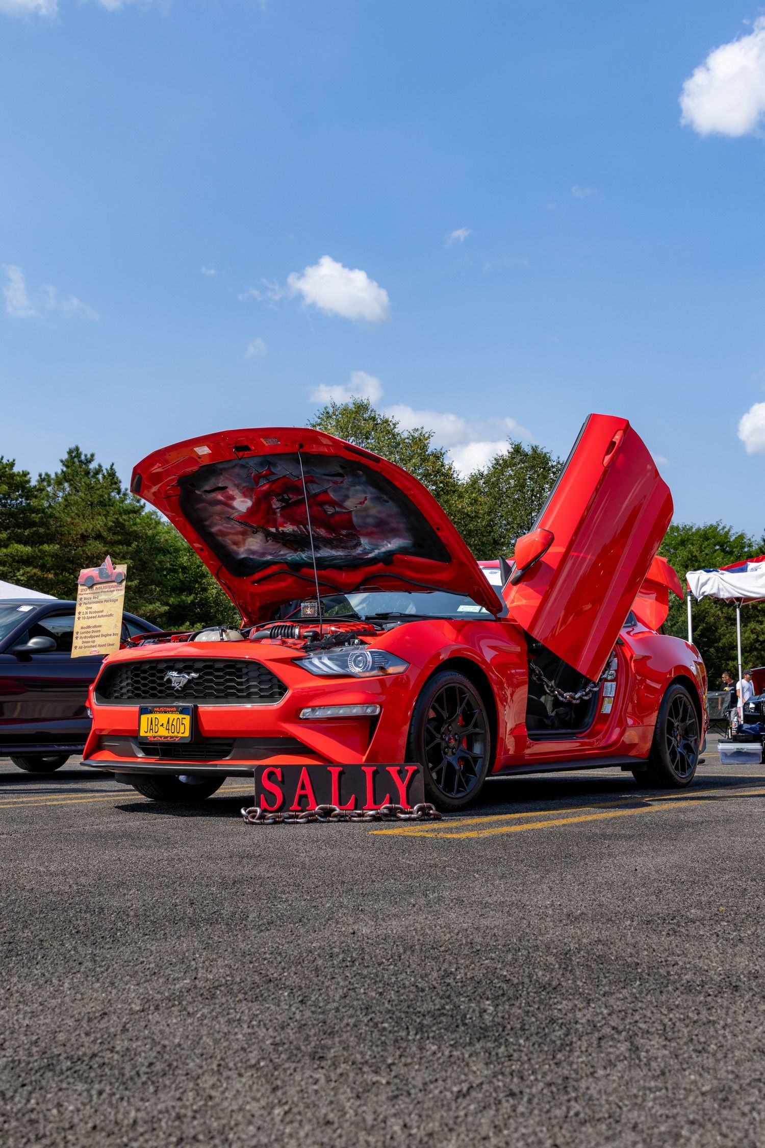 Red sports car with dramatic open scissor doors on display at an outdoor car show on a sunny day.