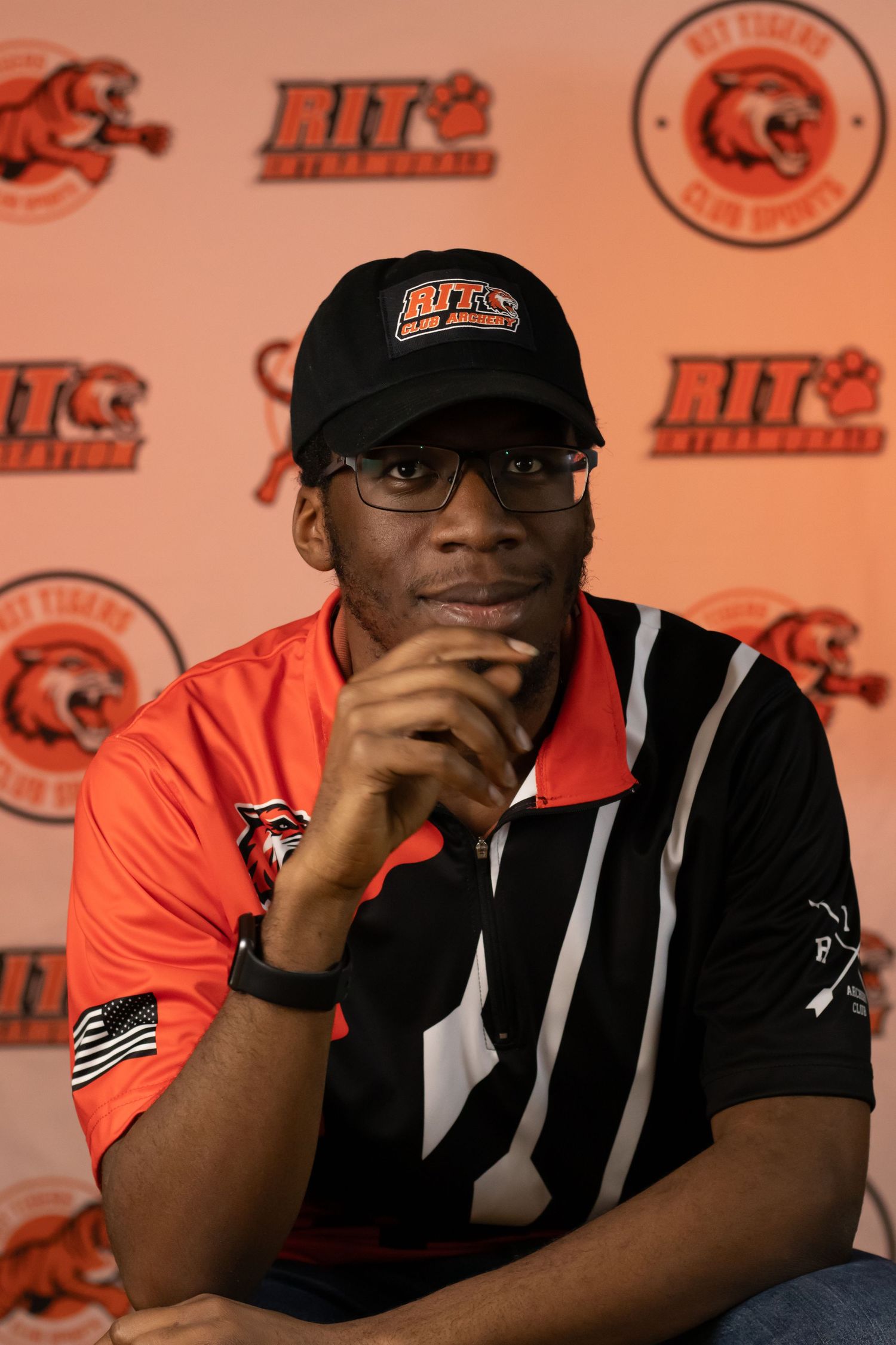 RIT athletics member in black cap and orange uniform poses thoughtfully against branded backdrop.