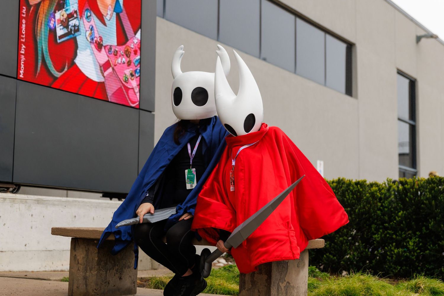 Two cosplayers dressed as Hollow Knight characters sit on a bench outdoors at a gaming convention.