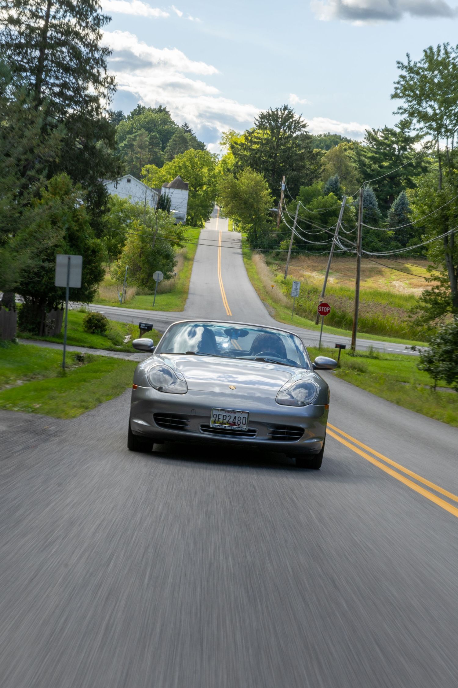 A silver Porsche 911 drives along a scenic country road lined with trees and power lines on a sunny day.