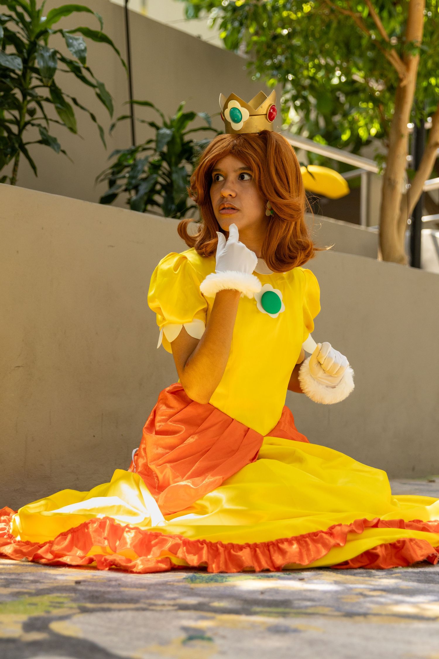 A cosplayer in a bright yellow and orange princess-style dress poses gracefully on the ground in an outdoor setting.