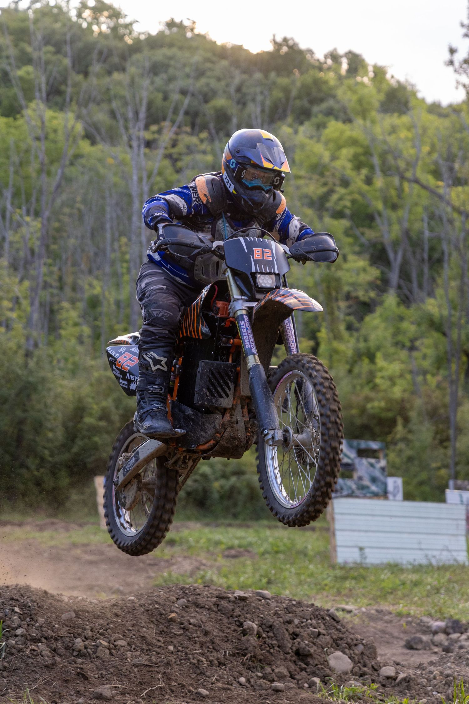 A sequence of motocross dirt bike jumps showing a rider performing aerial maneuvers over dirt mounds in a wooded area.