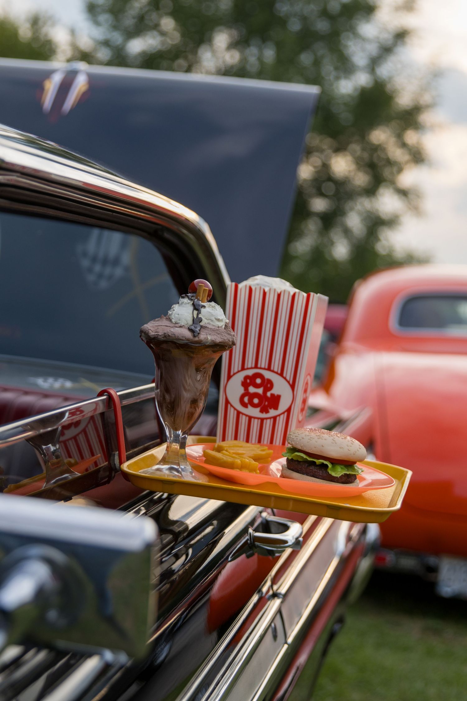 Vintage car drive-in movie theater tray with retro striped popcorn boxes and food service at dusk.