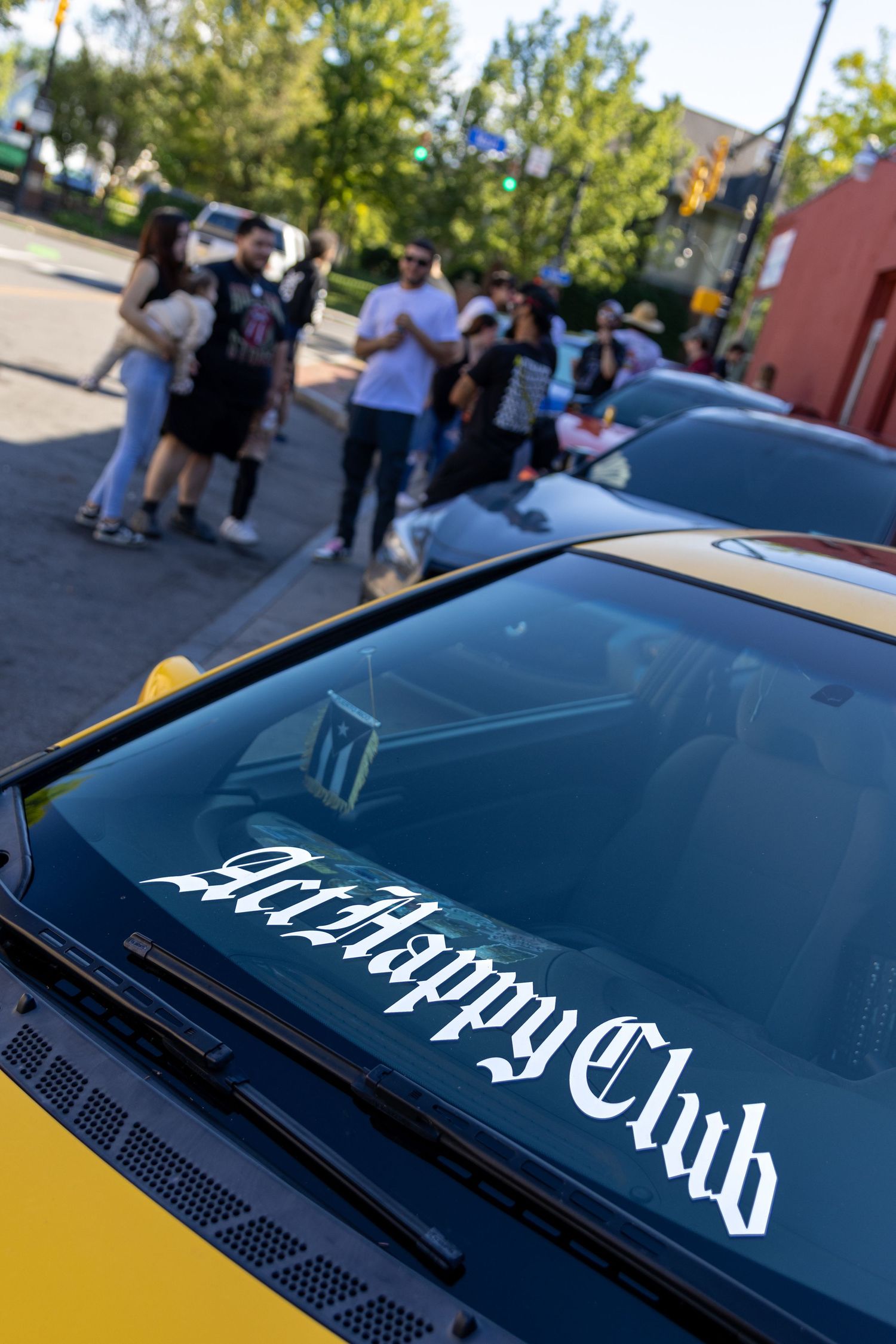 Yellow car with Midnight Club decal on windshield parked with group of people gathered in background.
