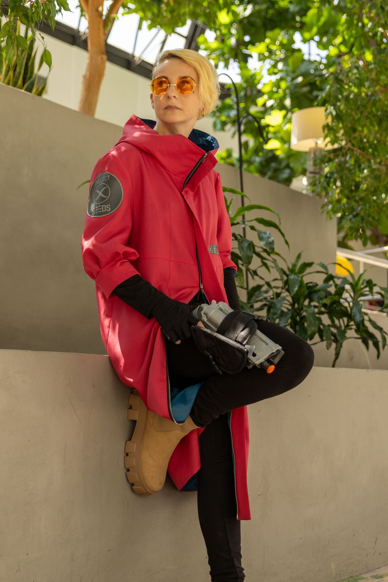 Cosplayer in pink jacket and black pants strikes dynamic pose holding prop weapon amid green foliage.