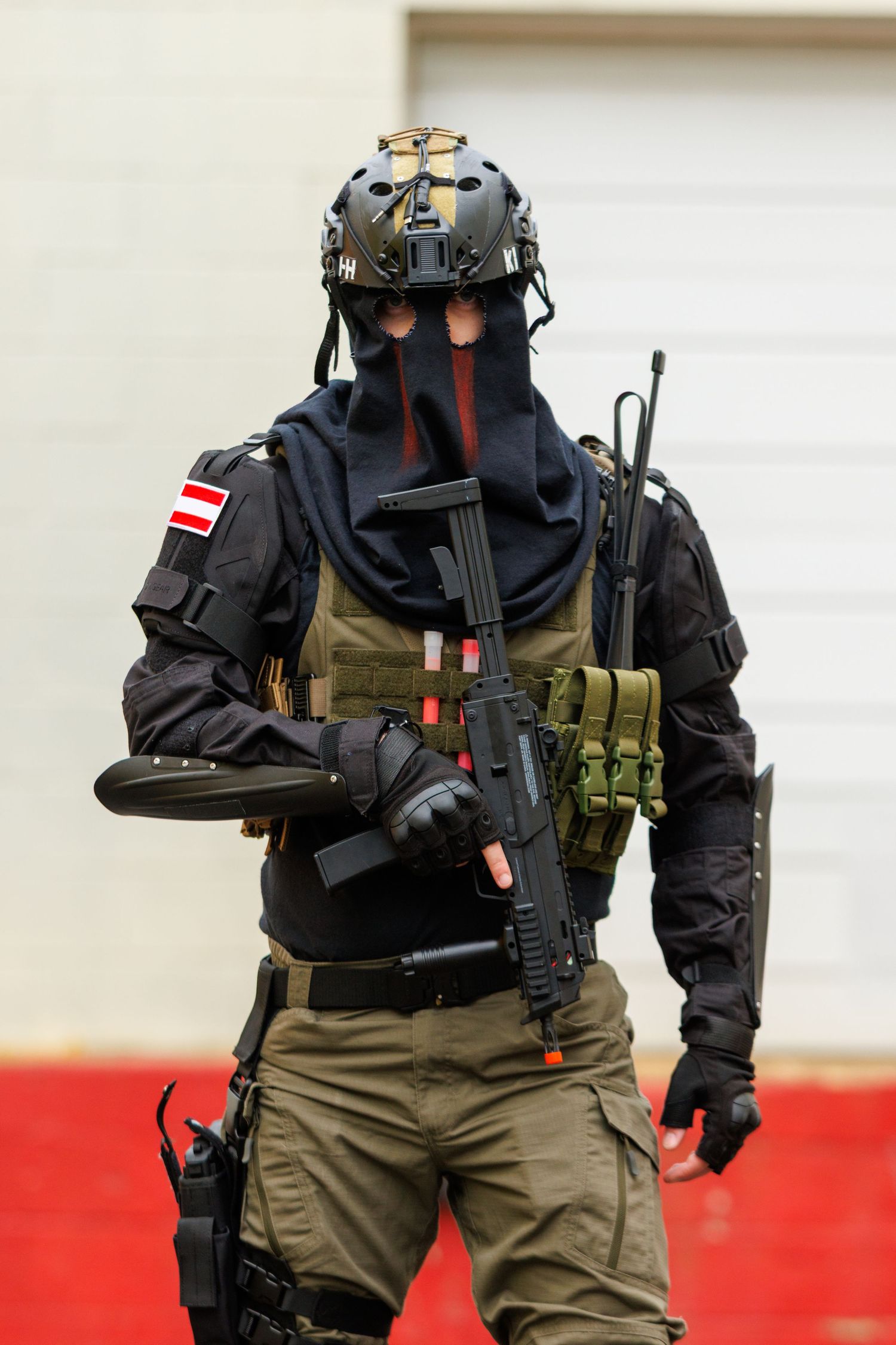 Person in tactical gear with helmet, face covering, plate carrier vest and rifle standing in front of a red and white wall.