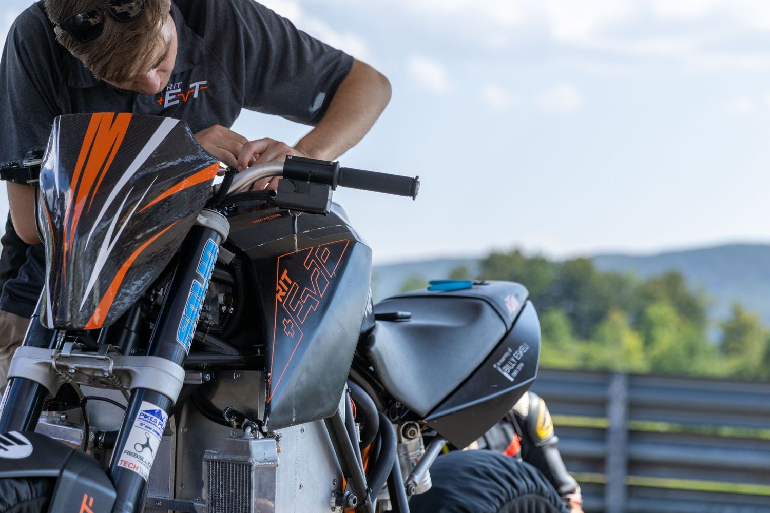 A motorcycle rider leans over a sleek black and orange sports bike against a scenic mountain backdrop.