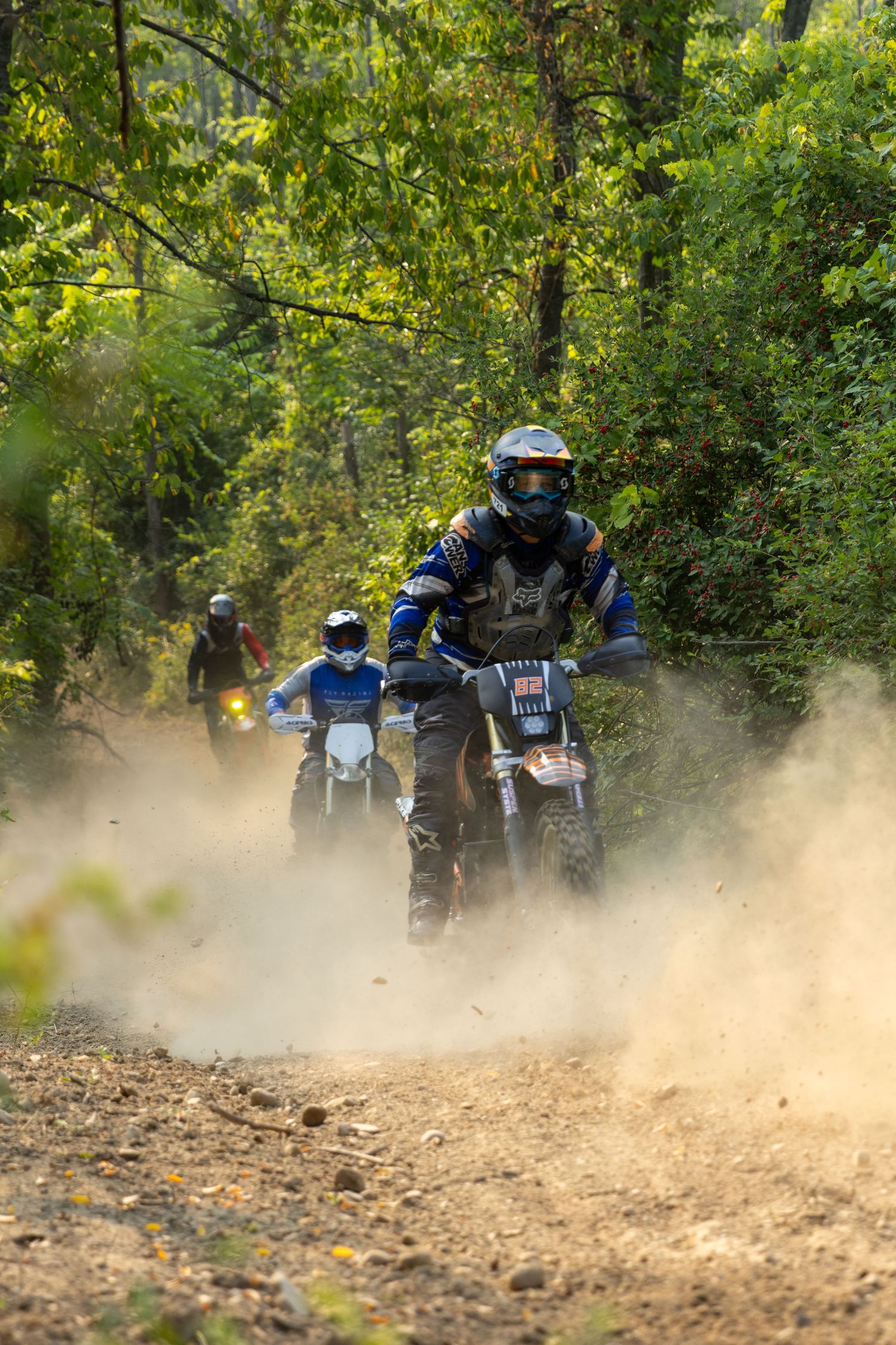 Multiple dirt bike riders kick up dust while riding in a line through a wooded area on a dirt trail.