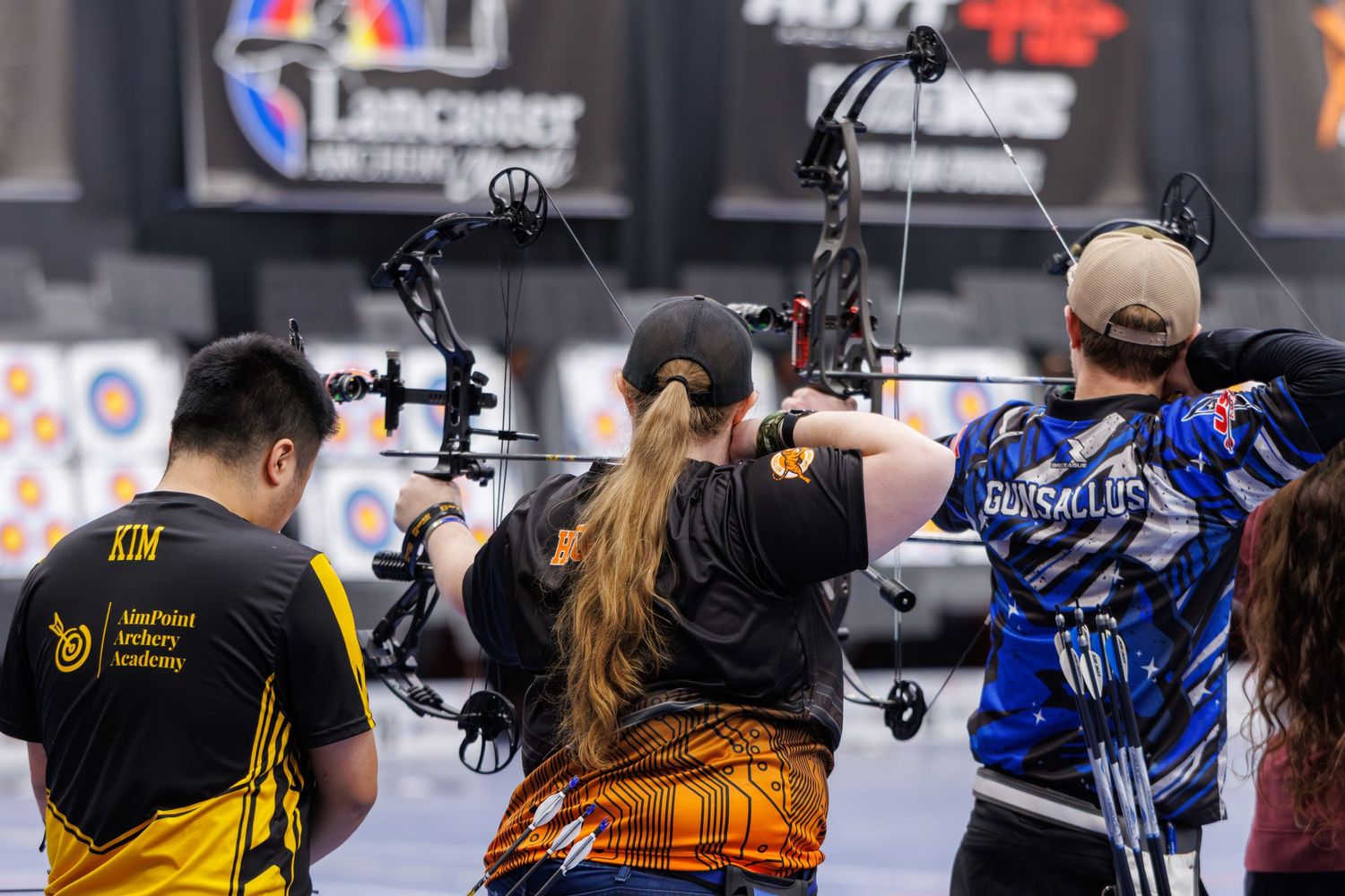 Three archers aiming compound bows at targets during an indoor archery competition.