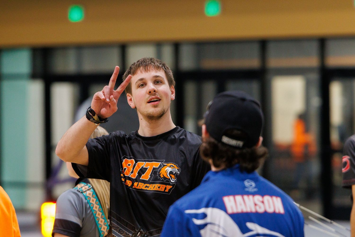 Archer in black BTC archery shirt giving peace sign at outdoor archery event.