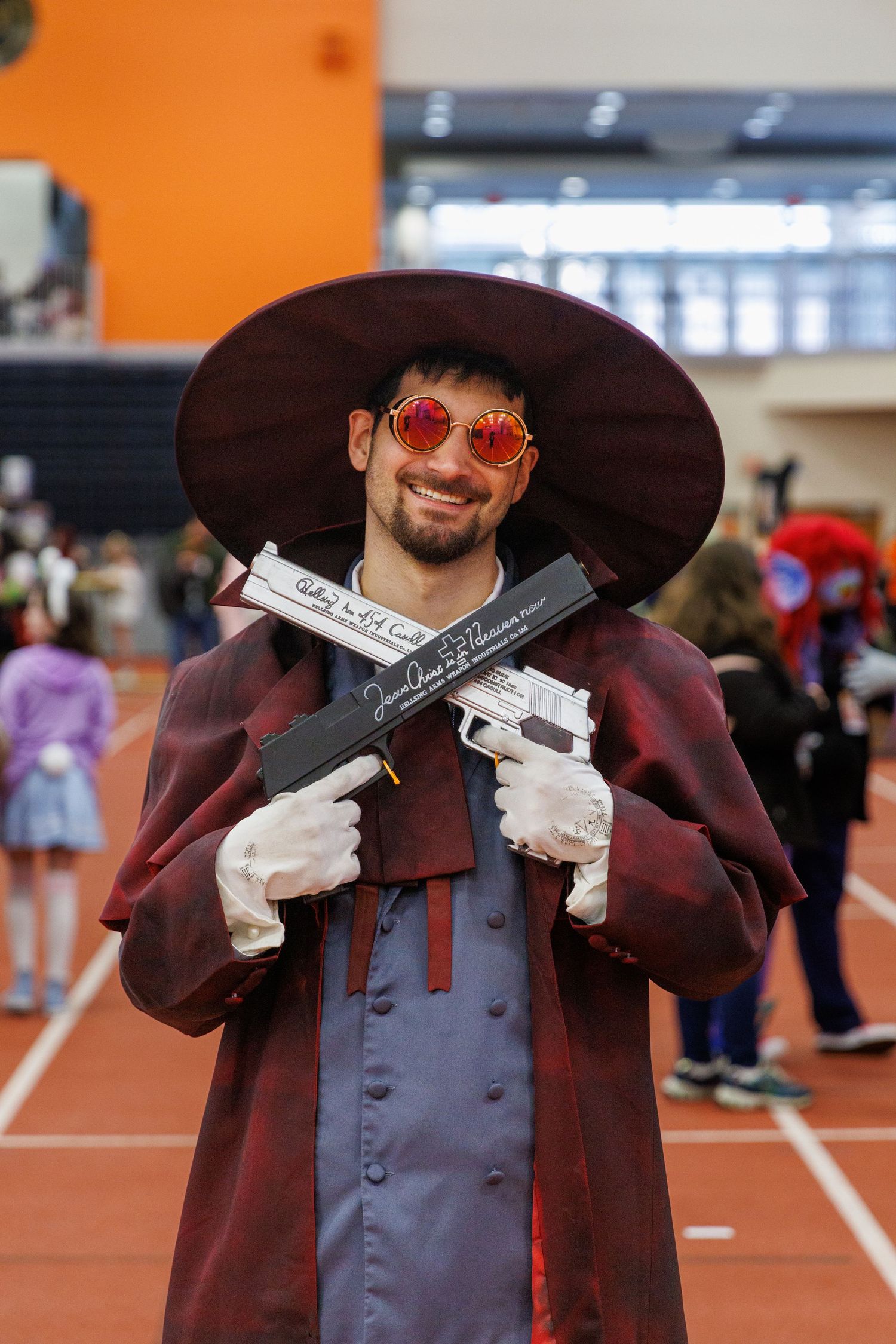 Man cosplaying in dark red coat, wide-brim hat, red sunglasses, white gloves, holding two prop guns at a convention.