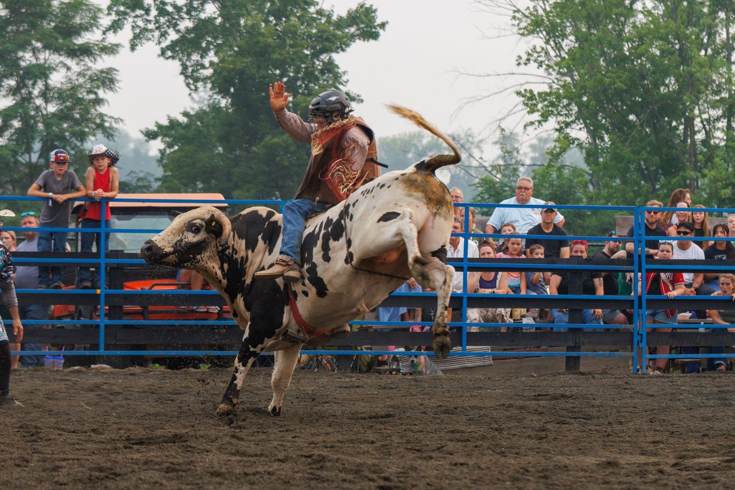A rodeo rider attempts to stay on a bucking bull at an outdoor arena with spectators watching behind blue rails.