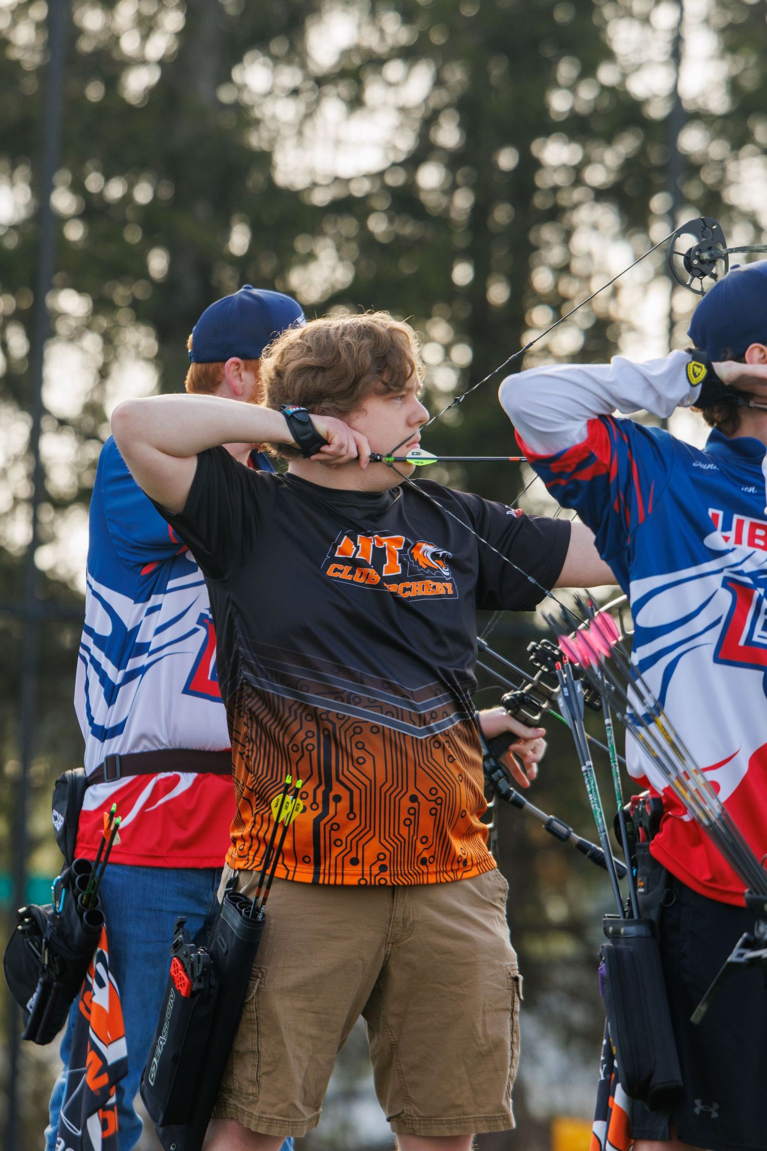 Three young archers drawing compound bows at an outdoor archery competition.