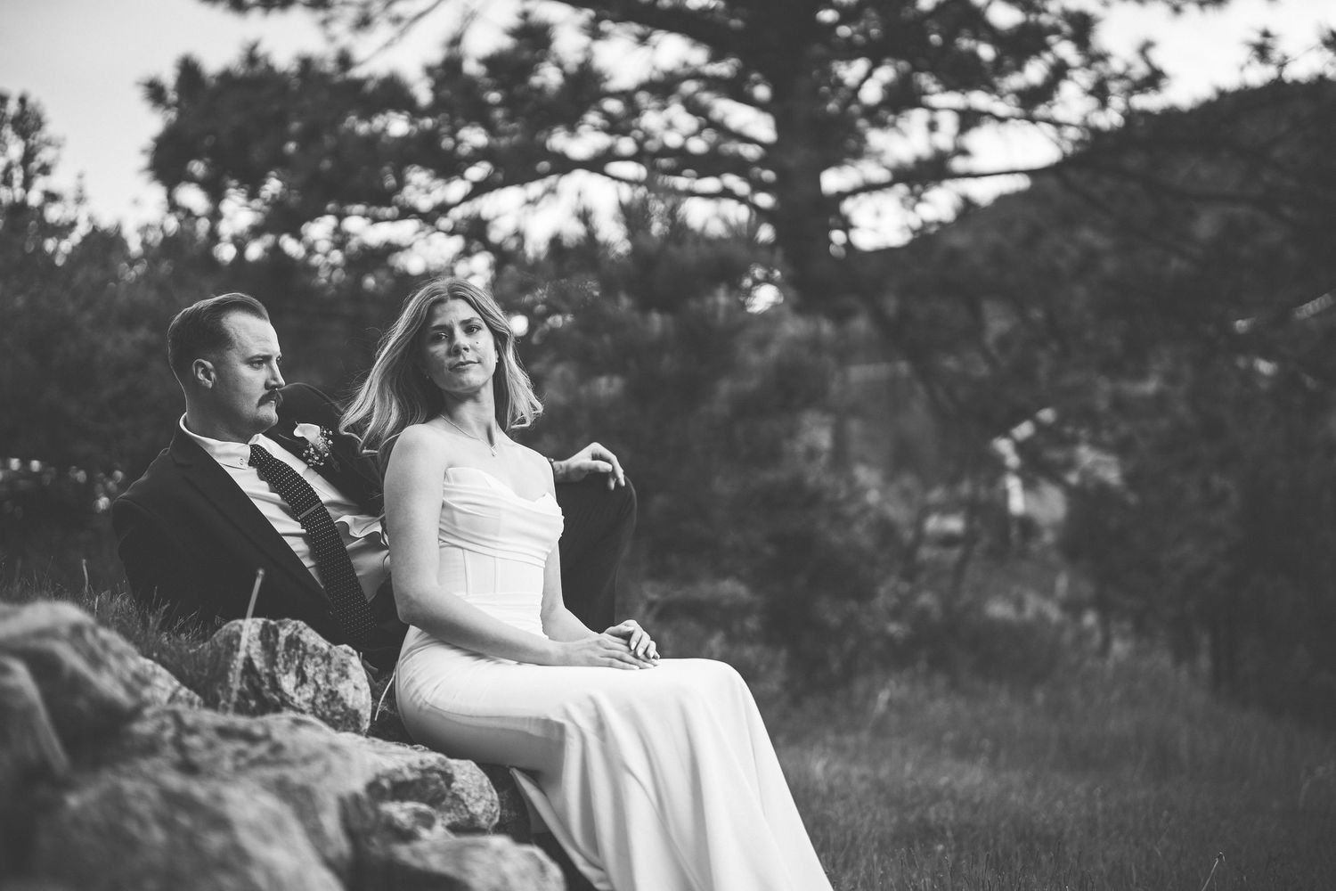A bride and groom share an intimate black and white moment while seated on rocky terrain outdoors.