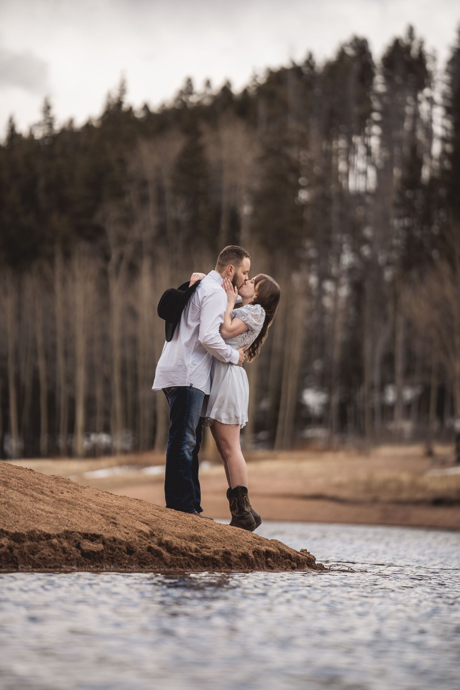 Couple shares romantic moment by lakeside with pine forest in background.