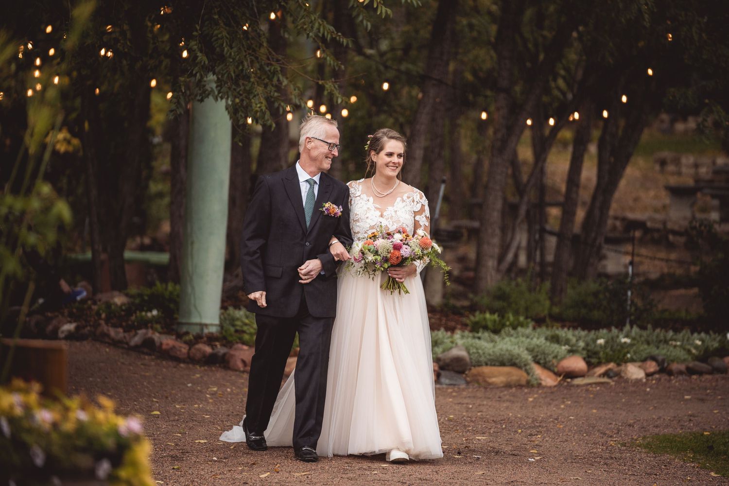 Wedding party walking down garden path illuminated by string lights at dusk.
