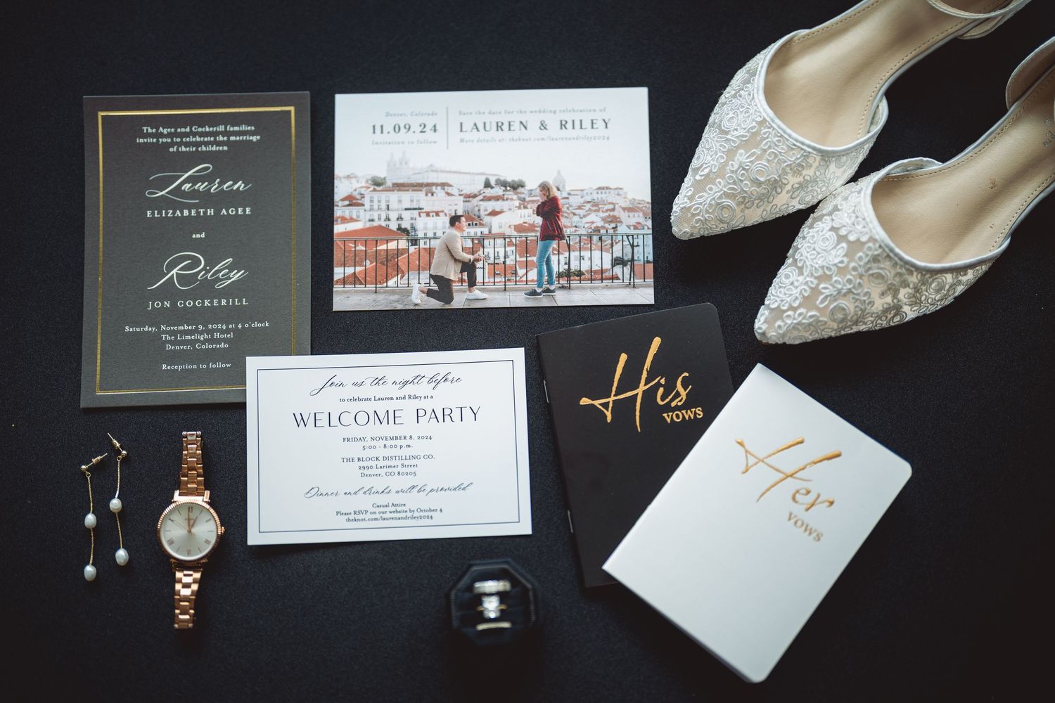 Wedding invitation suite and accessories laid out on a dark surface including invites and delicate shoes.