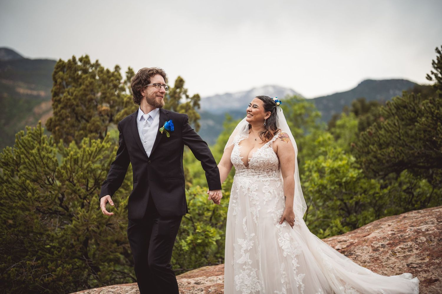 Bride in flowing lace gown and groom explore scenic mountain paths during their wedding photoshoot.