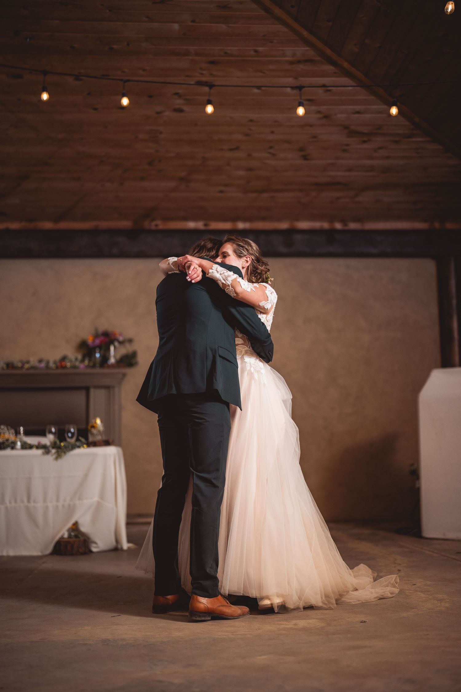 A couple shares an intimate slow dance moment in a rustic indoor venue with string lights overhead.