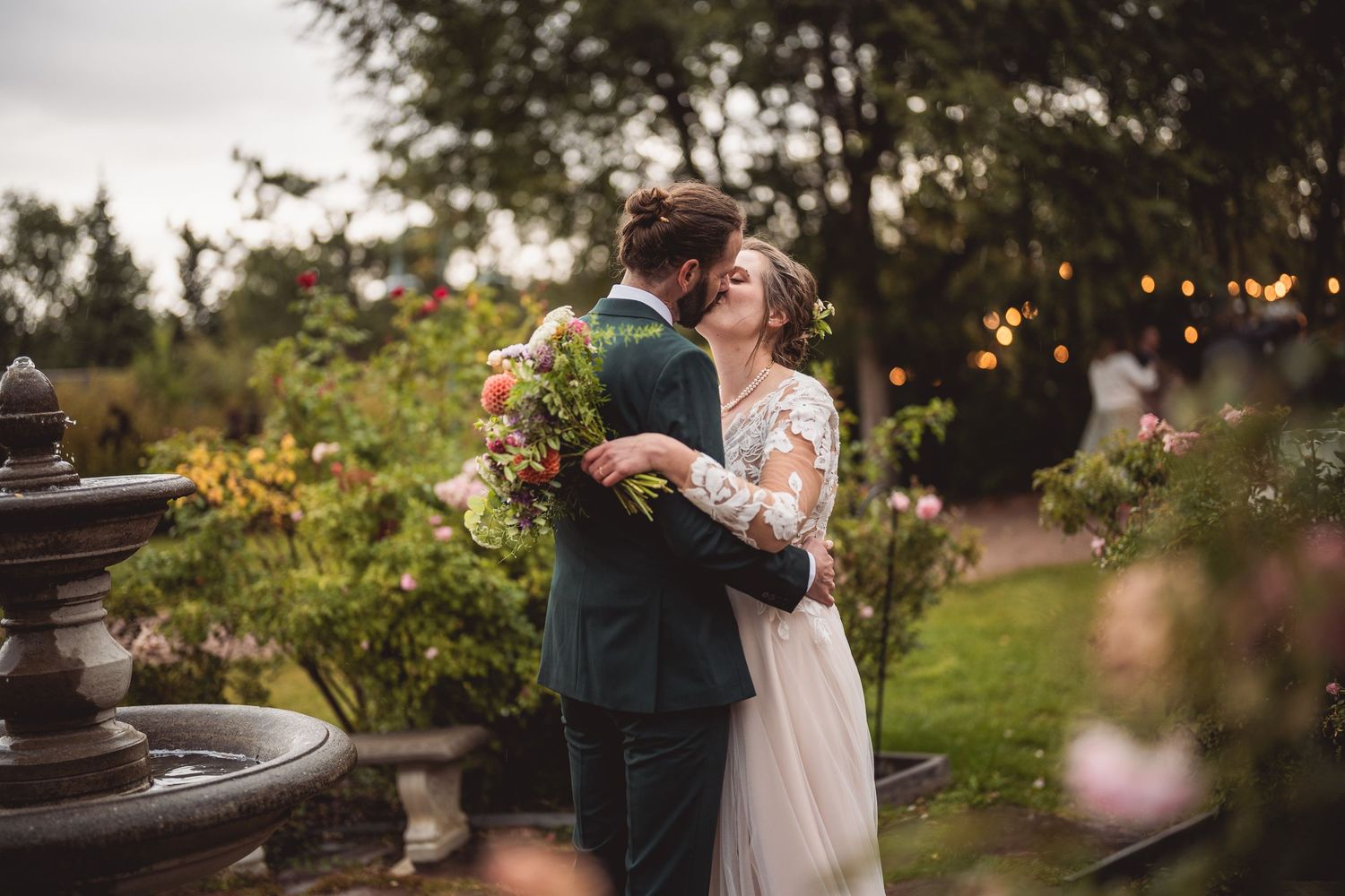 Newlyweds share an intimate embrace in a garden setting with string lights at dusk.