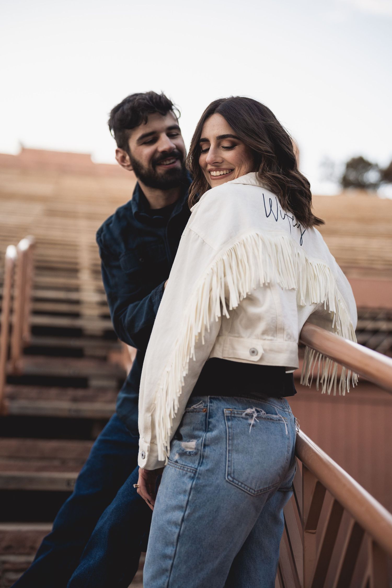 Casual couple in denim and fringe embracing on wooden stairs at sunset.