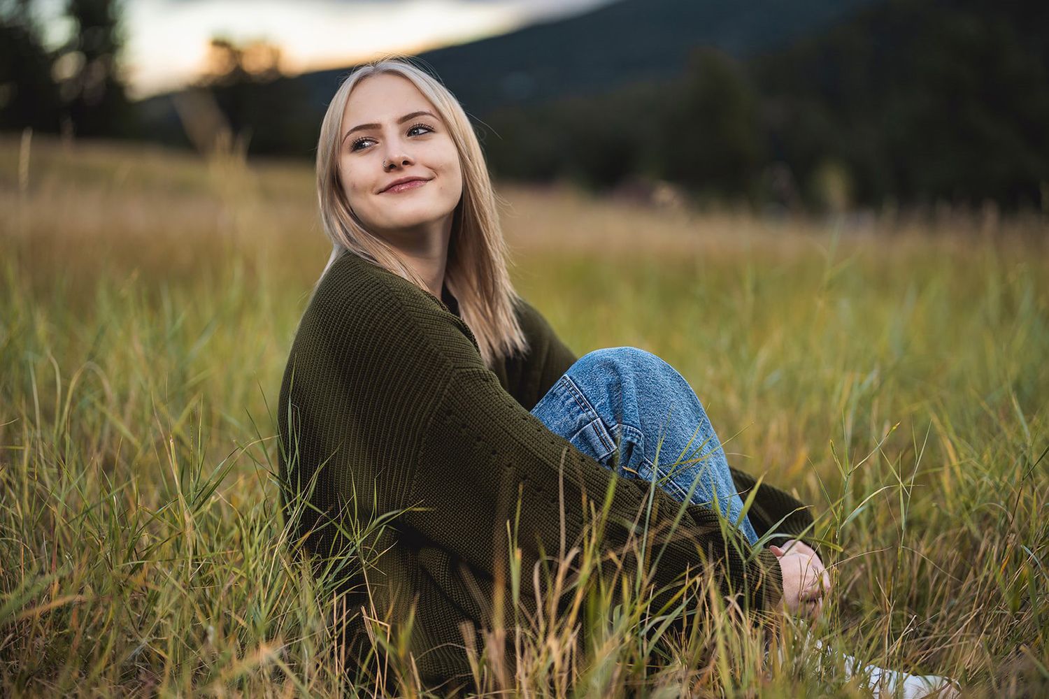 Smiling blonde girl in olive sweater and jeans sitting in a grassy mountain meadow at sunset.