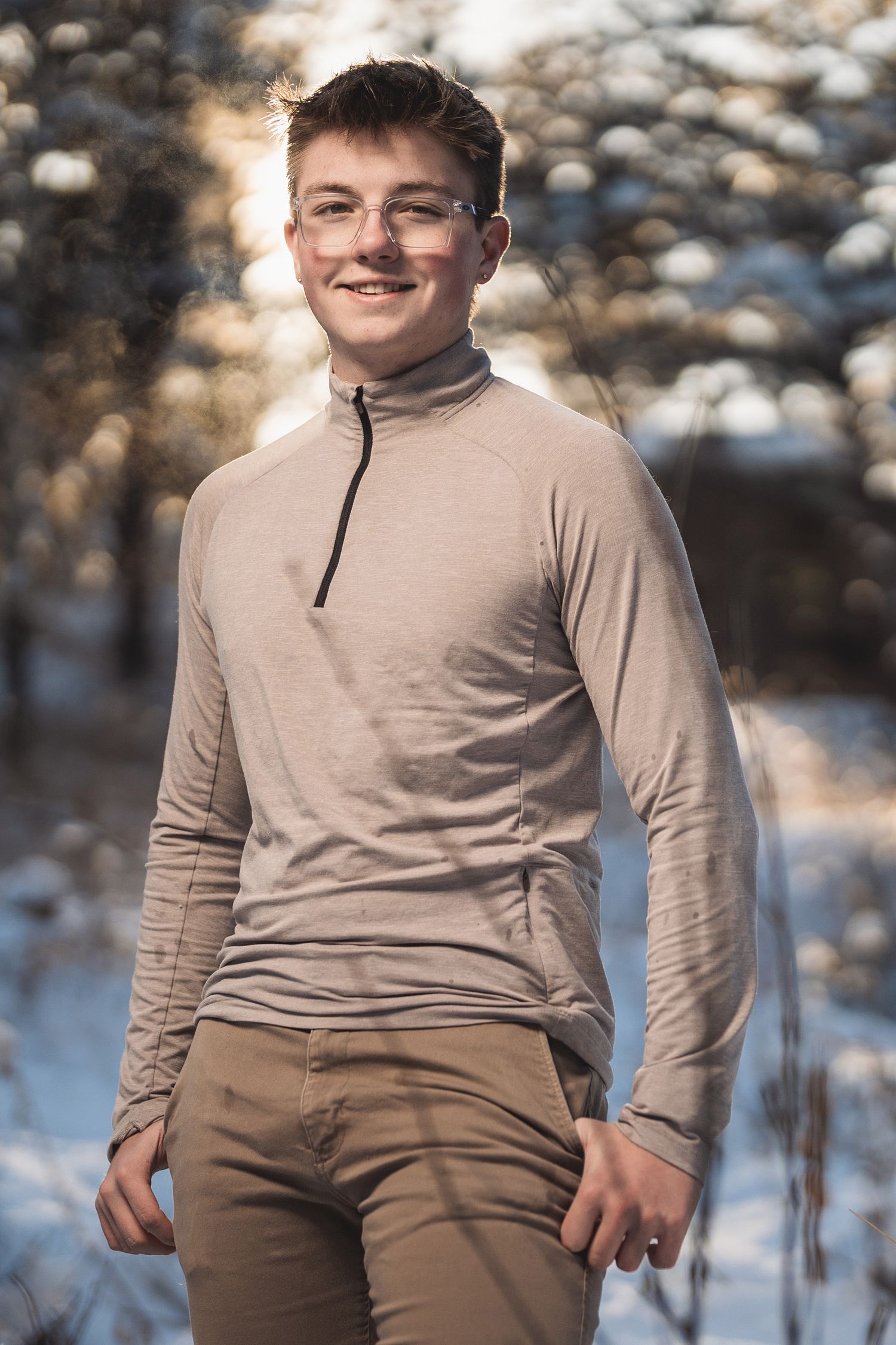 Young man in beige quarter-zip top and khaki pants smiling outdoors in a snowy winter forest setting.