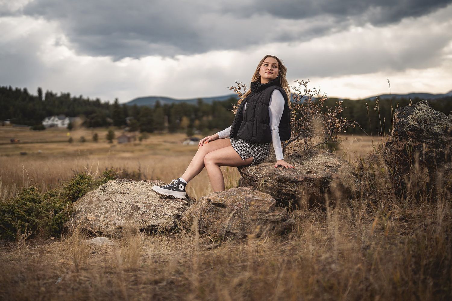 Young woman in plaid skirt sitting on rocks in a dramatic cloudy rural landscape.