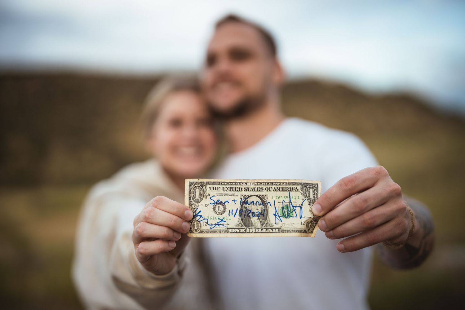 Couple smiling outdoors holding a decorated one dollar bill with the word LOVE written on it.