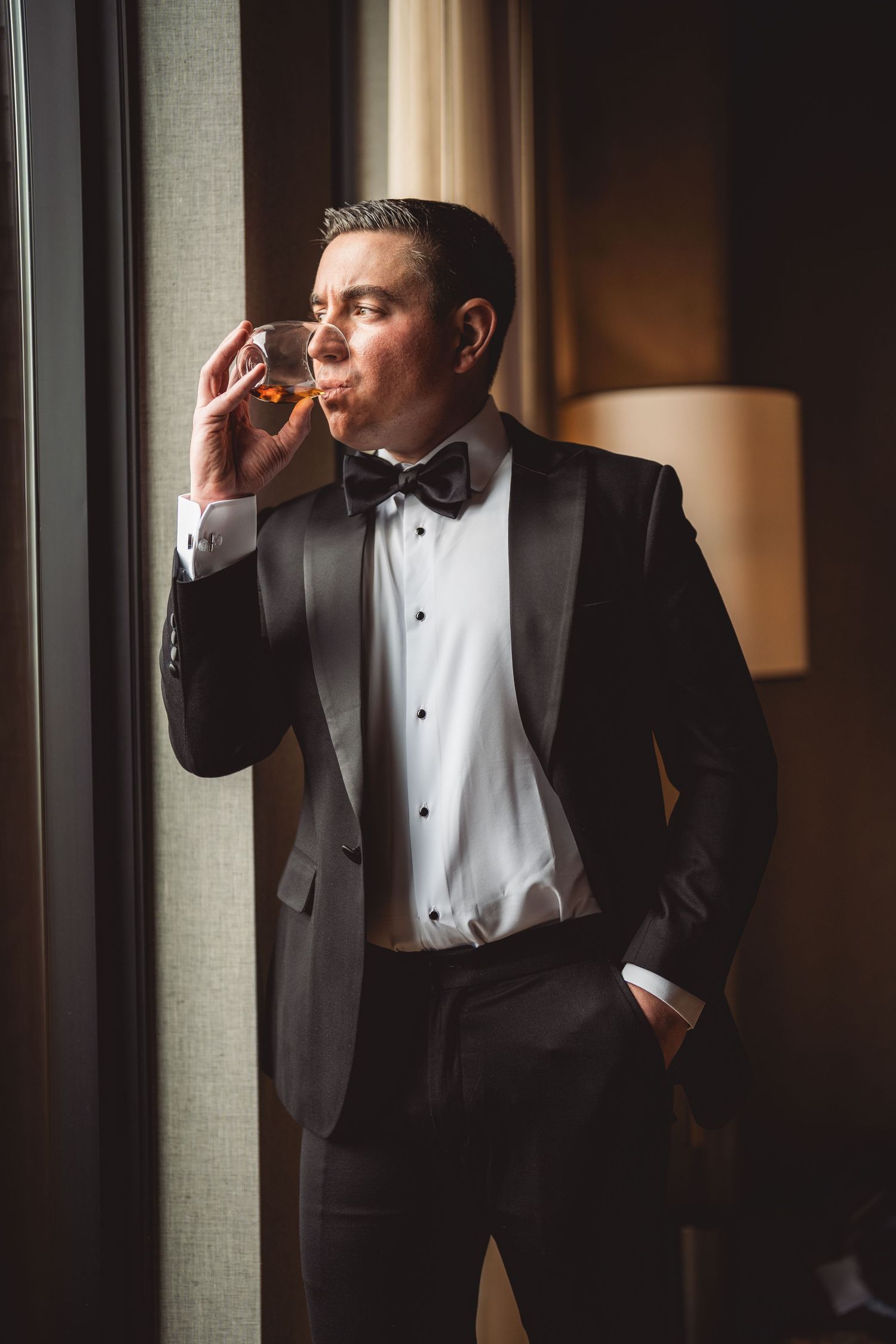 Dapper groom in black tuxedo and bow tie sips whiskey by a window before his wedding.