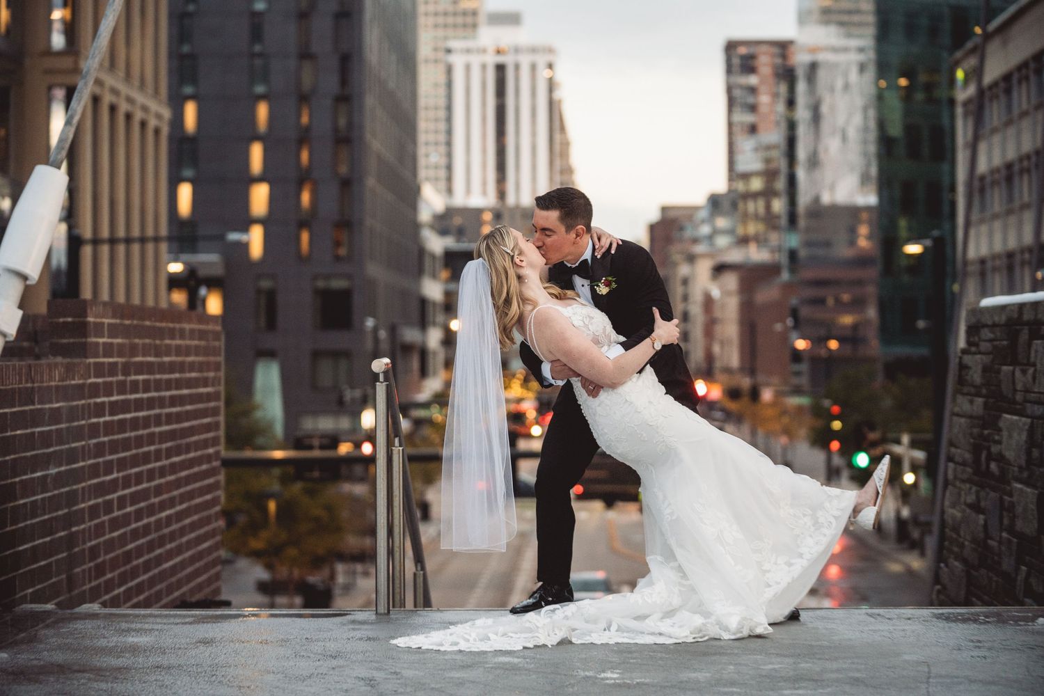 Bride and groom share a romantic dip kiss on a rooftop with a stunning city skyline backdrop at dusk.